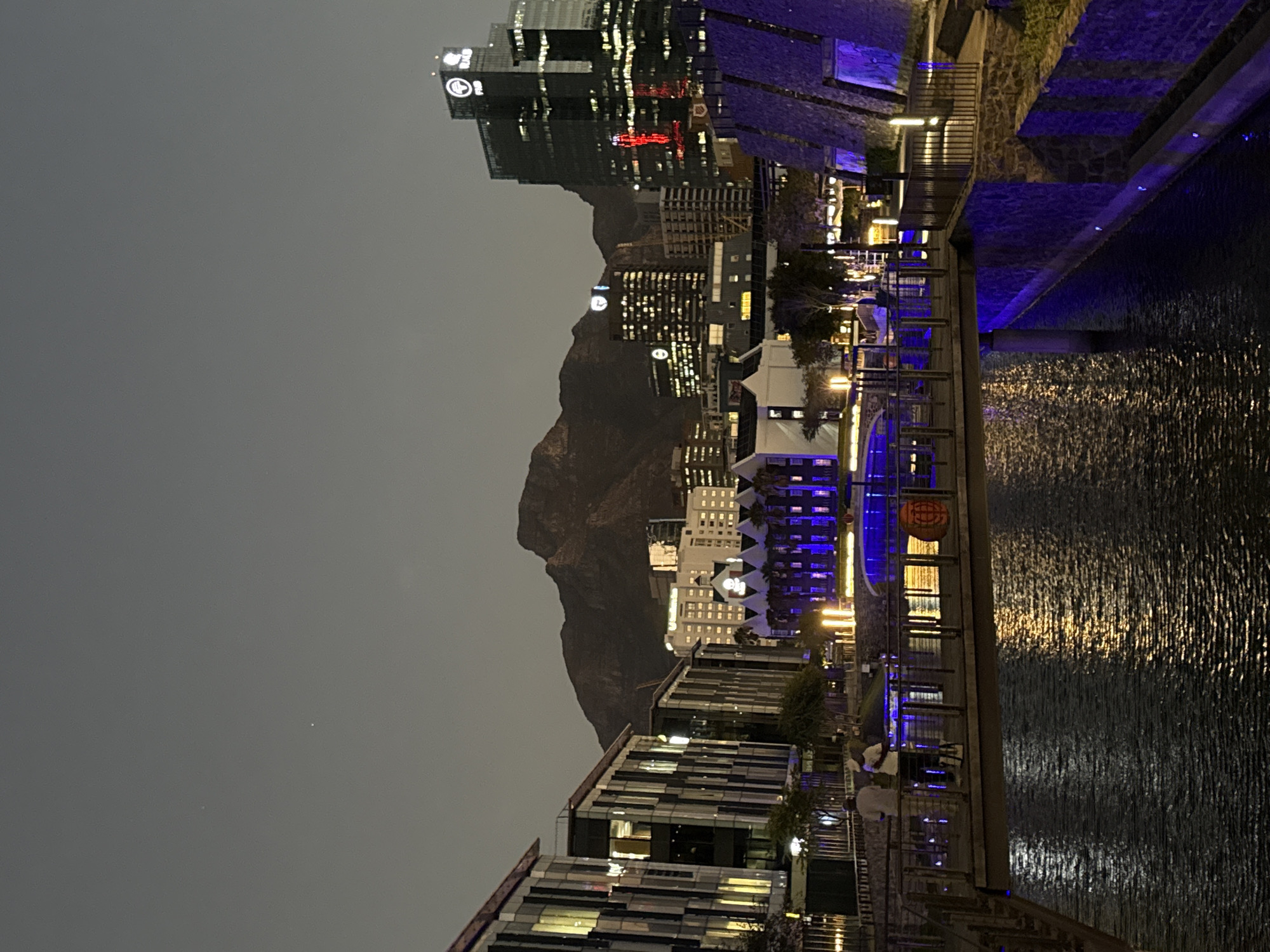 The image captures an evening scene in Cape Town, South Africa. In the background is the imposing outline of Table Mountain against a dusky sky. Below, there are modern buildings with illuminated windows, reflecting the city's skyline. Along the right side of the image is a body of water, possibly a canal or harbor, where reflections of nearby lights shimmer across the surface. A waterfront area is visible with boardwalk-style walkways lined with what appears to be restaurants or shops, indicated by the presence of outdoor seating and lighting fixtures. The scene seems to be a vibrant urban setting at night.