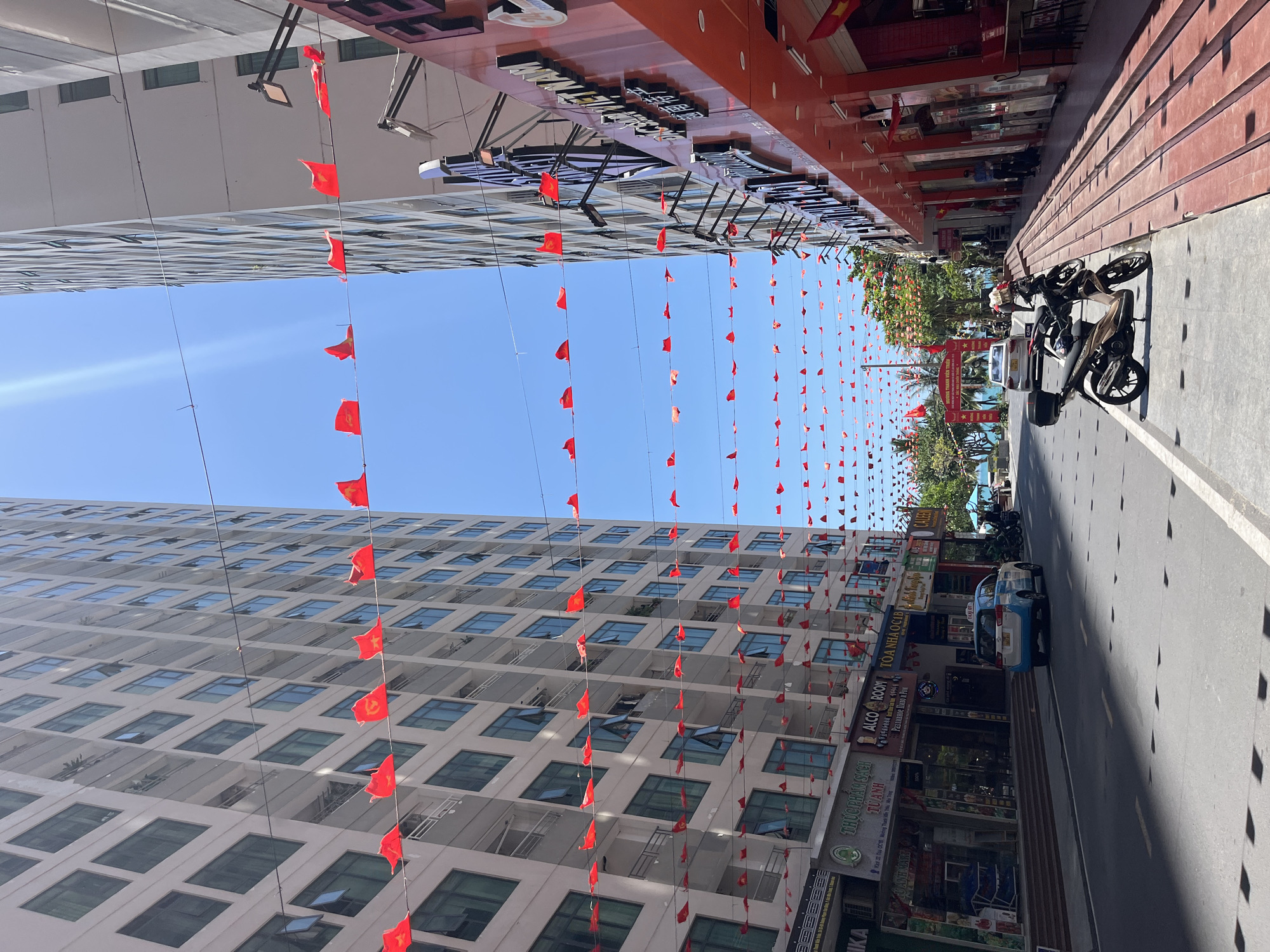 The image captures a street scene in Bac Nha Trang Ward, Vietnam. Red flags are strung across the street between buildings. The left side of the image features a tall, white building with multiple windows. The right side shows commercial buildings with various shops, including one labeled "ALCO ROOM" and another with a pharmacy sign, "THUOC PHAM SẠCH TU ANH". A white and blue taxi with the name "TAXI OT" is on the right. Parked motorbikes are present alongside the road. The sky is clear and blue. The photo is taken from a low angle.
