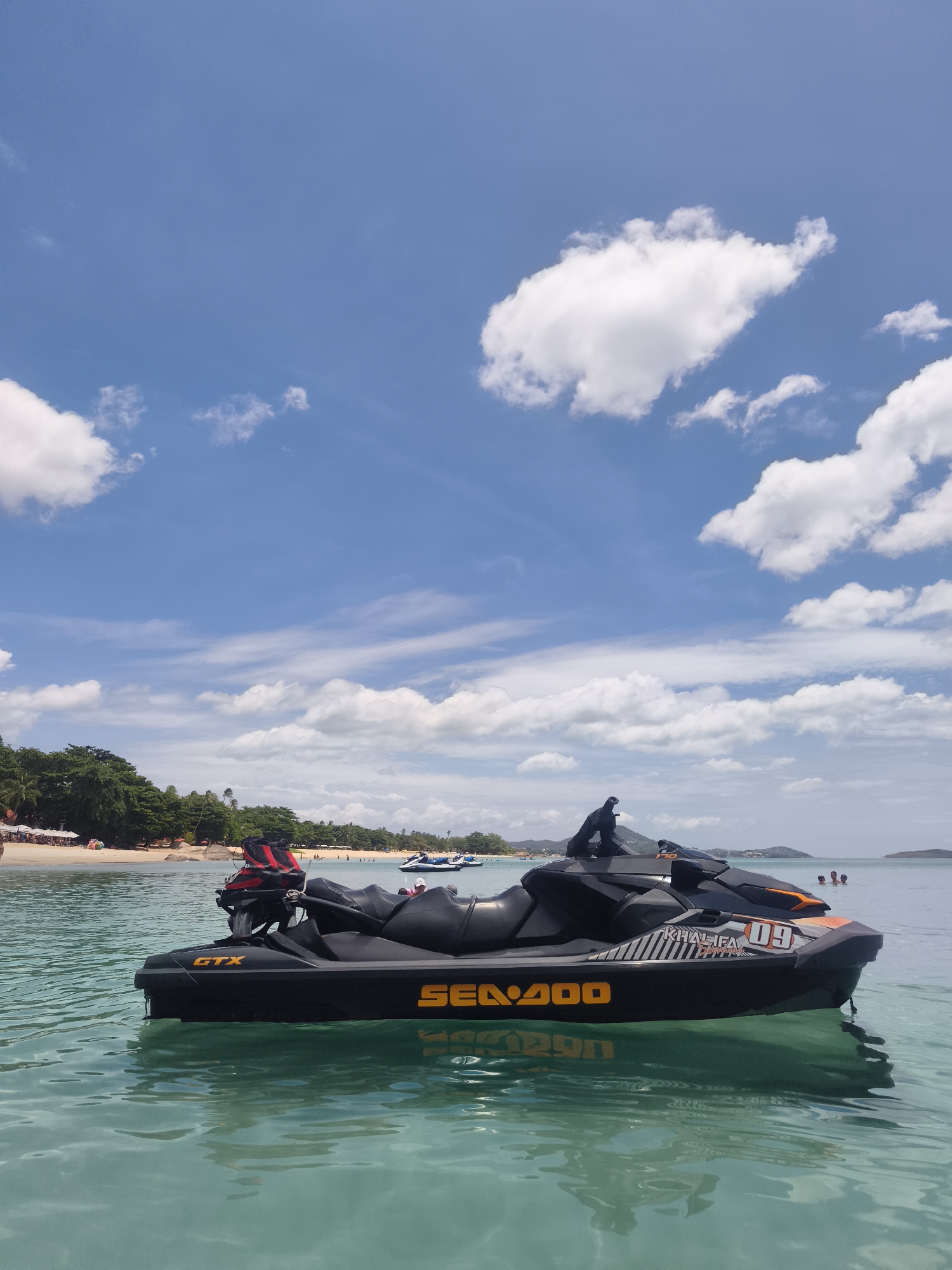 This is a ground-level, eye-level, medium shot photograph of a black Sea-Doo GTX jet ski floating on calm, clear turquoise water. The jet ski is positioned in the foreground, angled slightly to the right. It has a black body with orange accents and the "Sea-Doo" logo in orange lettering. A red and black life vest is visible on the back of the jet ski. The water has gentle ripples and reflects the blue sky and white clouds.
In the middle ground, a sandy beach is visible, lined with palm trees and vegetation. There are several beach umbrellas and lounge chairs on the beach, and a few people are scattered along the shoreline. Further out in the water, other jet skis and small boats can be seen, along with a few people swimming. The background consists of a hilly, tree-covered coastline and a clear blue sky with scattered white clouds. The location is Chaweng Beach, Ko Samui District, Thailand.