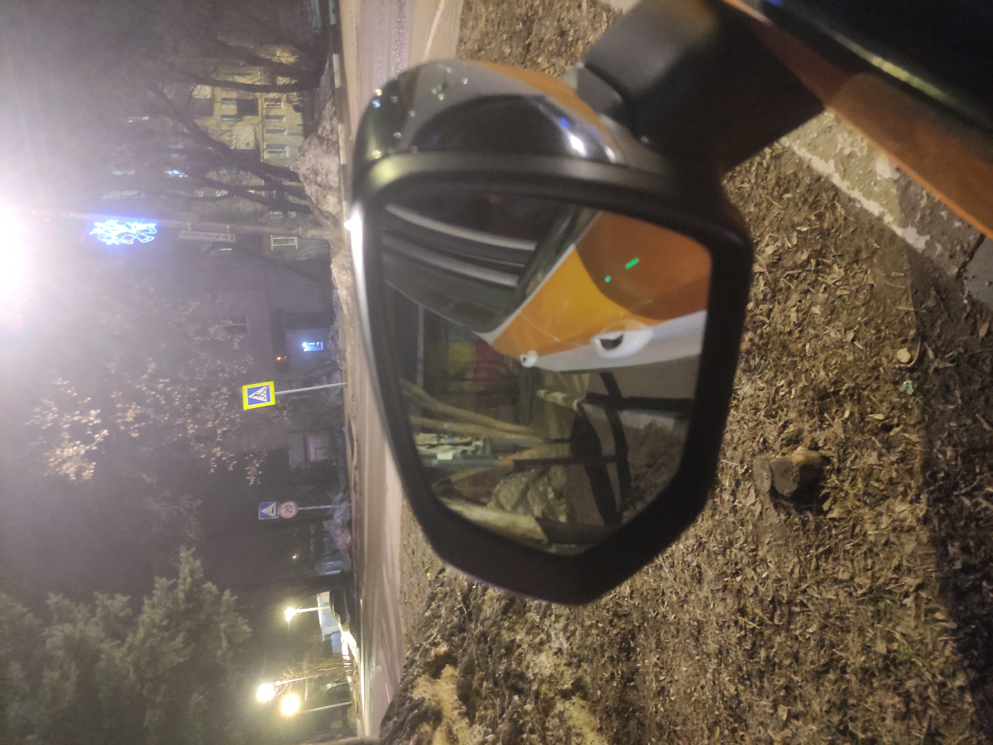 The image depicts a nighttime street scene in Kratovo, Russia, observed from a low perspective, likely from within or immediately adjacent to a parked vehicle. The foreground is dominated by a dark vehicle's side mirror, which reflects the two-tone orange/yellow and white side of the vehicle, along with a green indicator light and distorted elements of the surrounding environment. Small, bright specks, possibly water droplets, are visible on the mirror's upper surface.
To the left of the mirror, the ground is covered with dry, brown leaves and detritus, with a small, dark rock-like object resting on it. Dirty, partially melted snow is present on the ground to the lower right, adjacent to a faint curb line.
Beyond the mirror and foreground, a wet or damp paved road surface is visible. Along the roadside, several piles of dirty, compacted snow suggest winter or recent snowfall conditions. Various trees, some coniferous and others deciduous with bare branches, line the street. Multiple bright streetlights illuminate the scene, creating lens flare.
Traffic signs are visible: a circular white sign with a red border and the number "20" (indicating a speed limit) is stacked above a blue triangular sign with an unclear white icon. To its right, a prominent yellow square sign with a black border contains a white triangle with a black pedestrian symbol, indicating a pedestrian crossing. In the background, multi-story buildings with illuminated windows are faintly visible, and a bright blue, intricate decorative light (resembling a snowflake or star) is attached to a pole. No individuals are present in the scene.