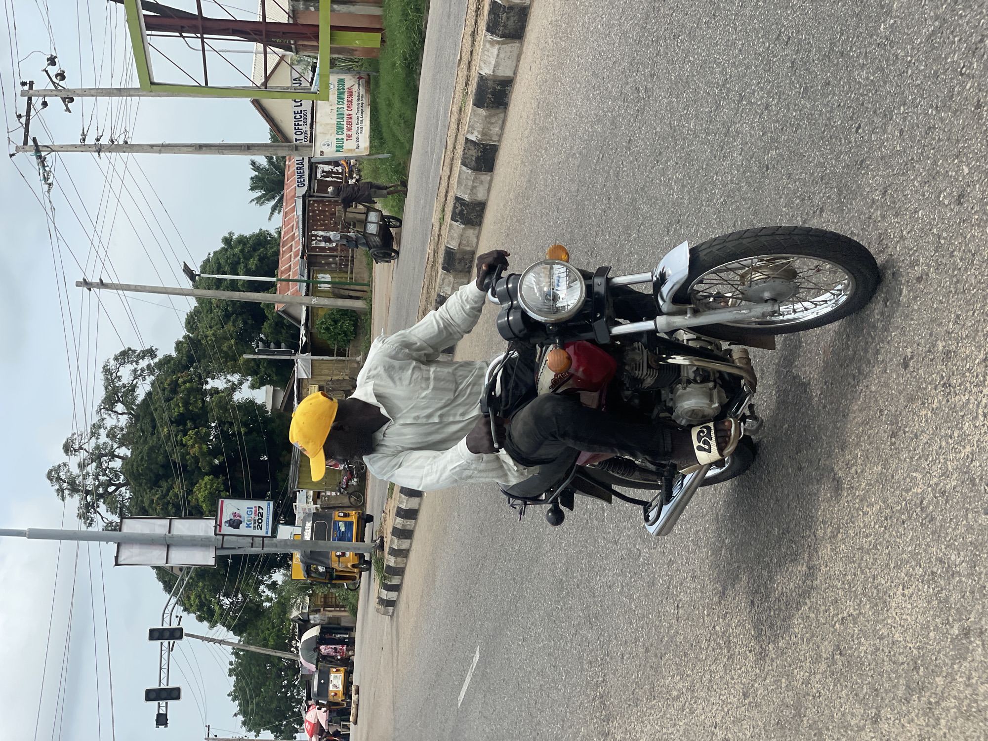 A man wearing a yellow baseball cap and a white striped button-down shirt rides a red motorcycle with a chrome exhaust pipe and handlebars. He is wearing dark pants and white flip-flops with a black logo. The motorcycle has a circular headlight and turn signals. The man is in motion on an asphalt road with a white lane marker. In the background, there are several yellow auto-rickshaws, some with people and umbrellas, parked along the side of the road. A traffic light and utility poles with wires are visible, along with trees and buildings. A green sign with white lettering reads "GENERAL T OFFICE LO JA" and another sign below it says "PUBLIC COMPLAINTS COMMISSION THE NIGERIAN OMBUDSMAN". The scene appears to be a street in Lokoja, Nigeria, with a cloudy sky overhead.