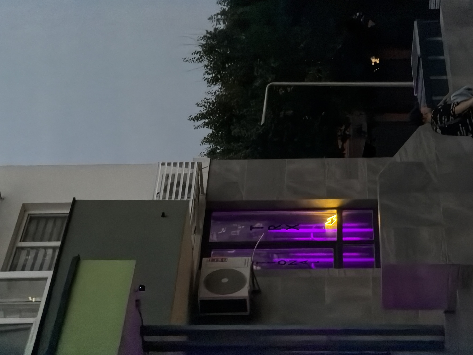 A night-time exterior shot captures a building with a green facade above a grey section. An air conditioning unit is mounted on the grey wall. To the right of the AC unit, a window is illuminated with vibrant purple light, and partially visible lettering in the window appears to spell out "TRX." A yellow neon sign in the shape of a "M" or a mirrored "W" is also visible within the purple-lit window. Below the window, a dark purple rectangular structure is present. Further to the right, a dark silhouette of trees is visible against a twilight sky. In the foreground, a person is seated, facing away from the camera, wearing a dark shirt with a patterned design and light-colored pants or shorts. They appear to be looking towards a dark, rectangular object. The scene is likely urban, given the building and street lighting.