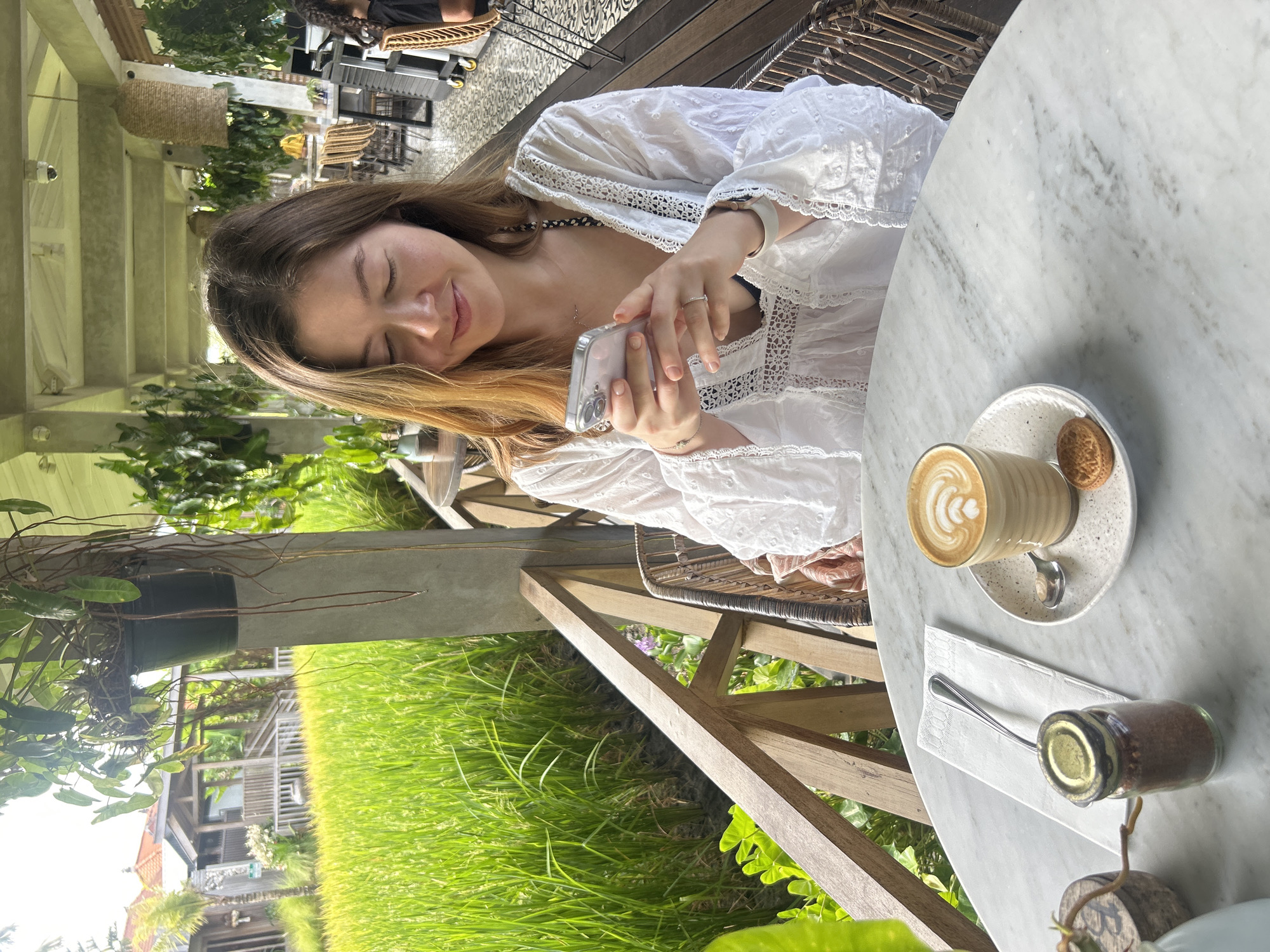 In North Kuta, Indonesia, a woman is seated at a marble table on a covered balcony overlooking a vibrant green rice paddy. She is wearing a white eyelet dress and is looking down at her phone, which she holds in both hands. A latte with foamy art sits on a plate with a cookie and a spoon in front of her. Next to the latte is a small glass jar containing brown granular sugar, with a spoon resting on a white napkin. A wicker chair is partially visible to her right. The balcony has concrete pillars and a wooden railing. In the background, other patrons are seated at tables, and a woman with curly hair is visible in the far right of the frame. Lush green plants hang from the ceiling and are scattered throughout the area. The rice paddy extends into the distance, with small buildings visible beyond it.
