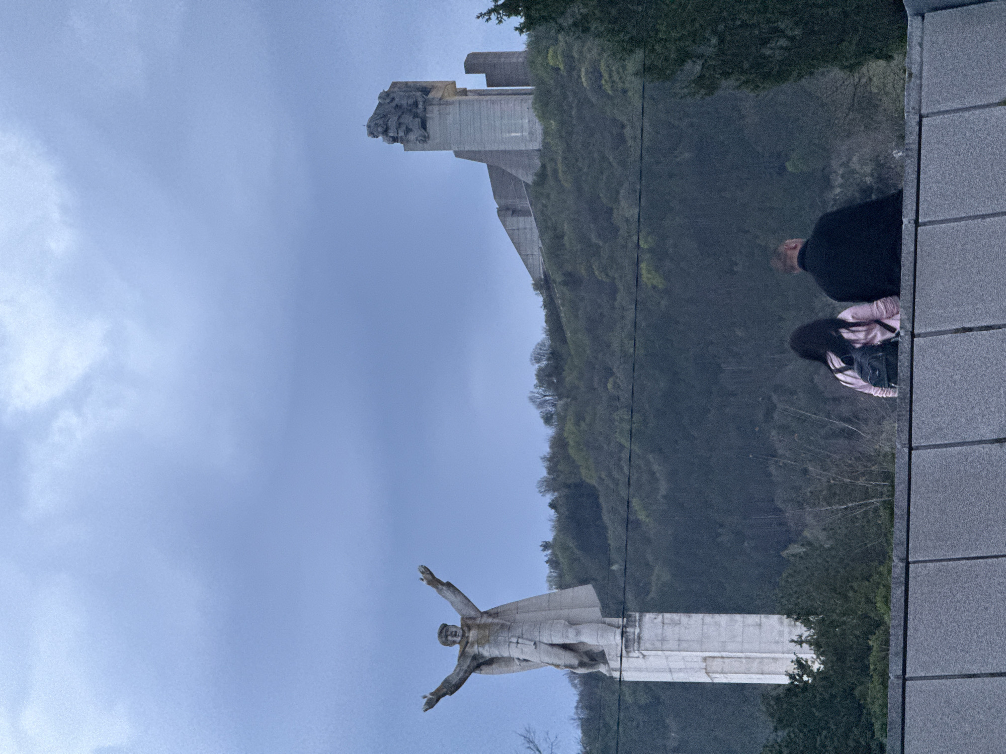 From a high vantage point in Shumen, Bulgaria, a photograph shows a panoramic view encompassing several landmarks. The Monument to 1300 Years of Bulgaria is positioned on a hill overlooking the city. Below this, another monument of an outstretched person is evident. Two individuals, one in dark clothing and one in lighter pink, are visible on a platform, possibly looking at the view. The sky is overcast, lending a muted tone to the scene. The surrounding landscape includes green foliage indicating the presence of trees and vegetation.