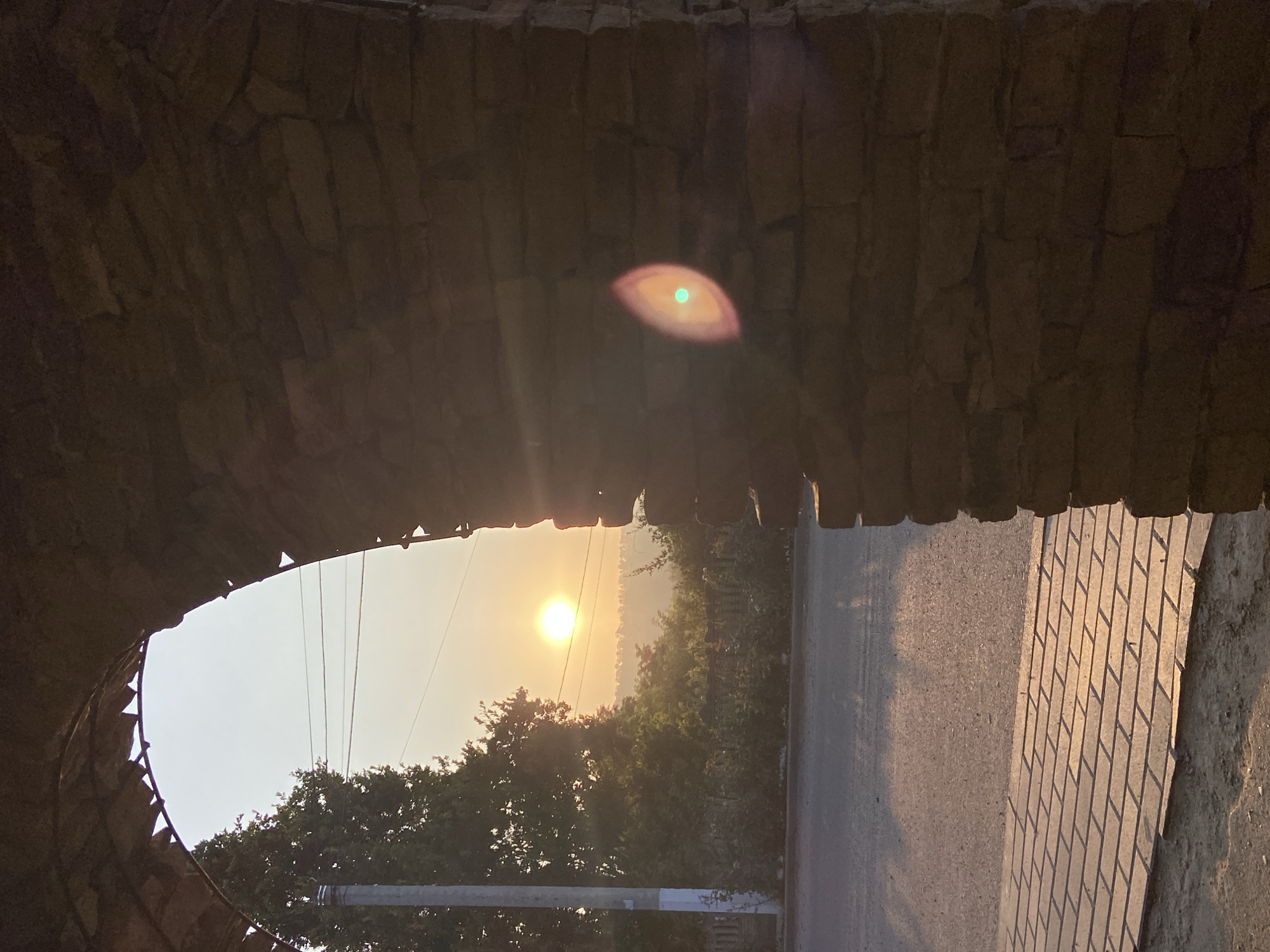 This is a vertical, eye-level, medium shot of an arched stone passageway with the sun setting in the distance. The stone archway frames the view of a rural landscape during sunset. The arch is constructed from roughly cut, irregular brown and grey stones, and it casts a shadow over the right side of the image. Through the arch, a bright, hazy sun is visible on the horizon, casting a warm glow over the sky, which transitions from a pale yellow near the sun to a light grey at the top of the frame. Below the sun, a line of trees and vegetation creates a dark silhouette. In the foreground, a paved pathway made of rectangular stones is illuminated by the setting sun, with long shadows stretching across it. To the left of the pathway is a paved road. A utility pole with wires is visible to the left of the road. There is a lens flare on the right side of the image, appearing as an oval shape with a bright green center. There are no people or animals visible in the image. The scene suggests an early morning or late evening setting in a quiet, possibly rural or suburban area.