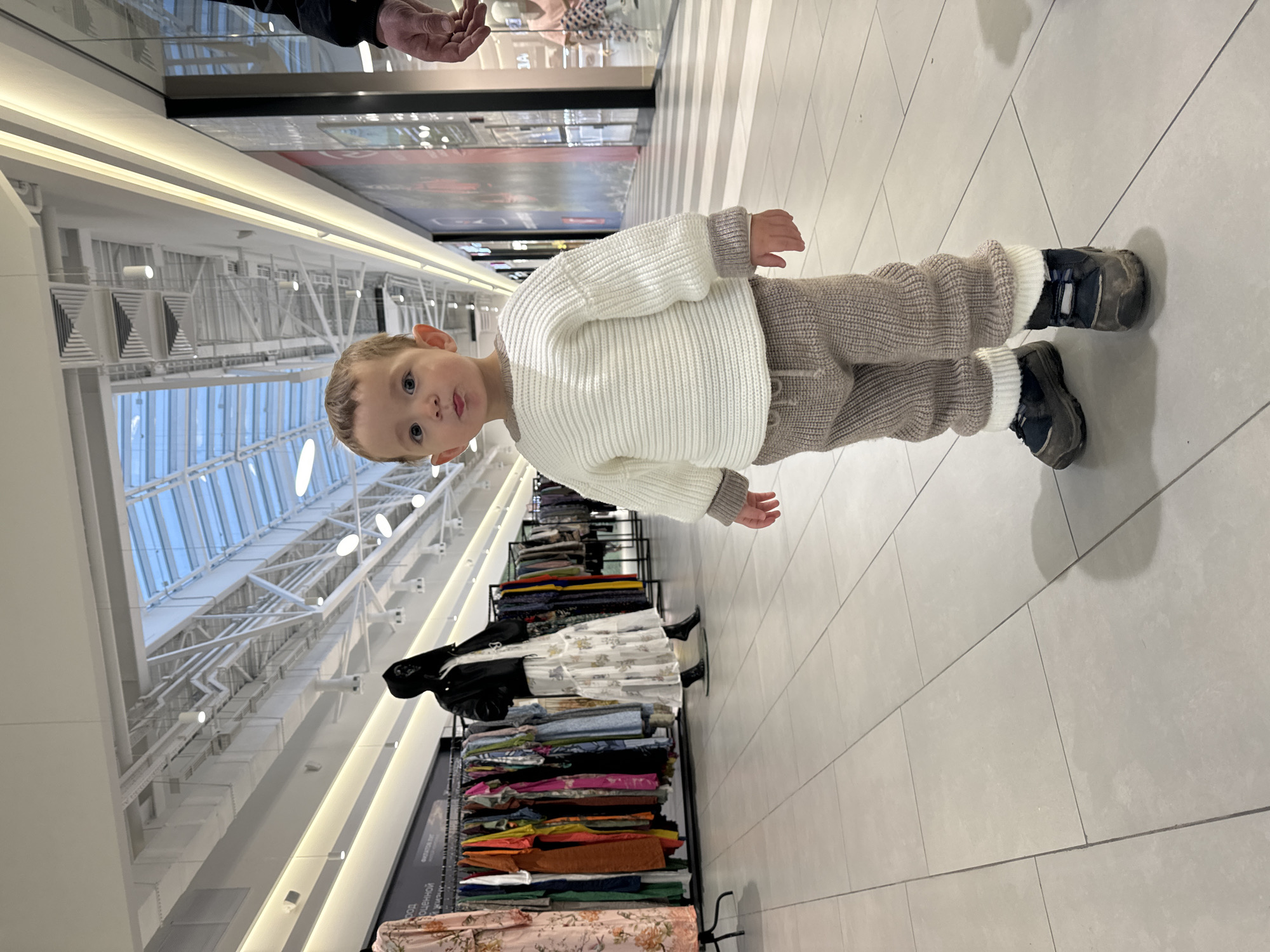 A young child in a white sweater and light brown pants stands on a light grey tiled floor. The child faces the camera and is centered in the frame. In the background, a rack of colorful fabrics and clothes is visible. The location appears to be an indoor shopping mall with a skylight. The architecture suggests a modern structure, potentially in Kriukovo, Russia.