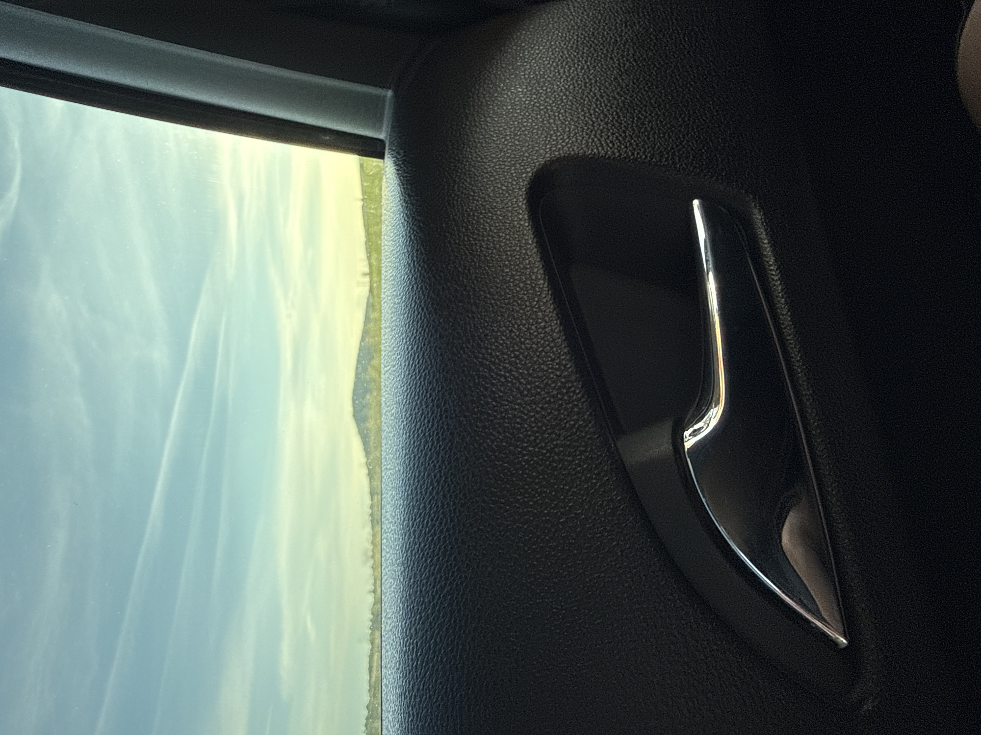 The image shows the interior of a car, specifically the door panel. The panel is black with a textured surface. A chrome door handle is visible. Through the car window, a landscape with a light blue sky and light green land is seen. The location is not Slavyani, Bulgaria.