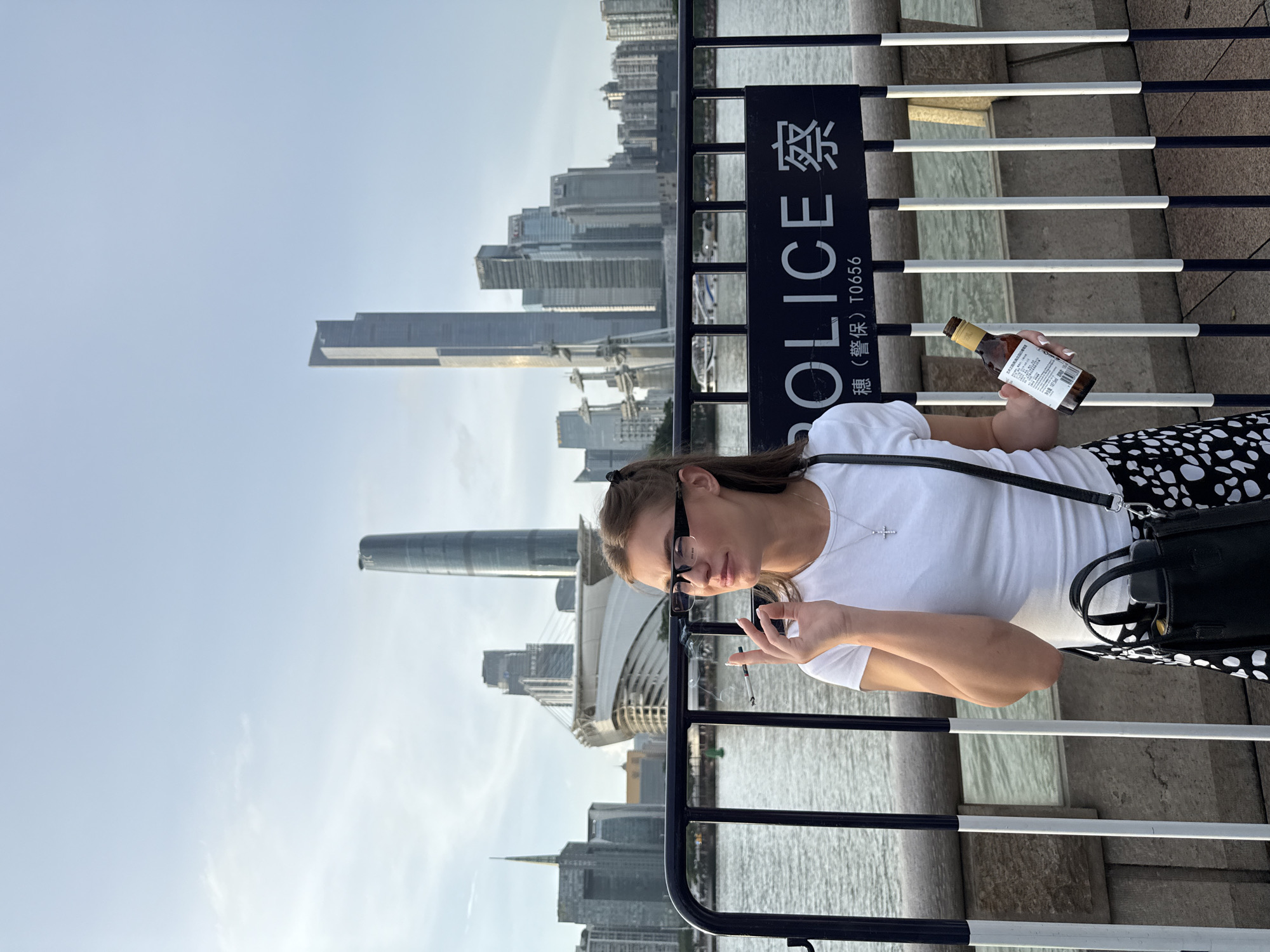 A young woman with brown hair, wearing glasses, a white t-shirt, and a black and white polka dot skirt, stands near a black metal fence. She is holding a small brown bottle in her right hand and a cigarette in her left hand, which she is smoking. She appears to be looking at the camera and has a relaxed expression. Behind her, a cityscape with tall buildings and a body of water are visible. A sign on the fence reads "POLICE" in large white letters, with Chinese characters above and below it. The time of day appears to be late afternoon or early evening, with a clear sky. The location is identified as Haizhu District, China.