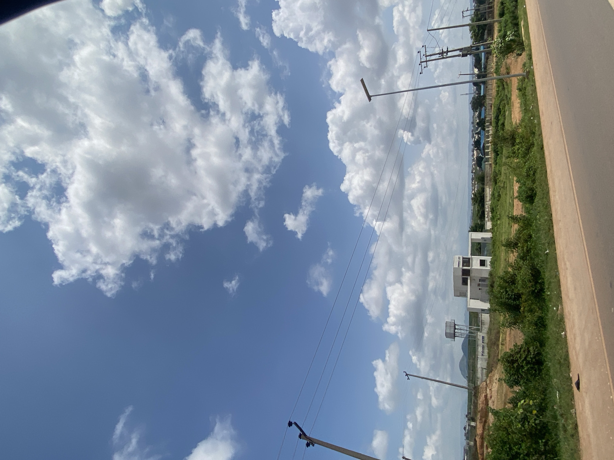 The image depicts a semi-arid landscape under a partly cloudy sky, with cumulus clouds scattered across a bright blue expanse. The foreground is dominated by a dirt road, and beyond it, a grassy embankment with scattered bushes and a lone sandal. In the mid-ground, a modern white building with a flat roof and visible windows stands beside a water tower. Further in the distance, power lines stretch across the scene, supported by utility poles. A row of houses with blue roofs is visible in the far background, along with some vegetation and what appears to be a mountain or large hill in the distance. The overall impression is one of a developing area on the outskirts of a city. The specific location within Ado, Nigeria, is not explicitly identifiable from the visual elements alone, but the landscape suggests a region with tropical savanna climate characteristics. There are no people or active interactions depicted in the image.