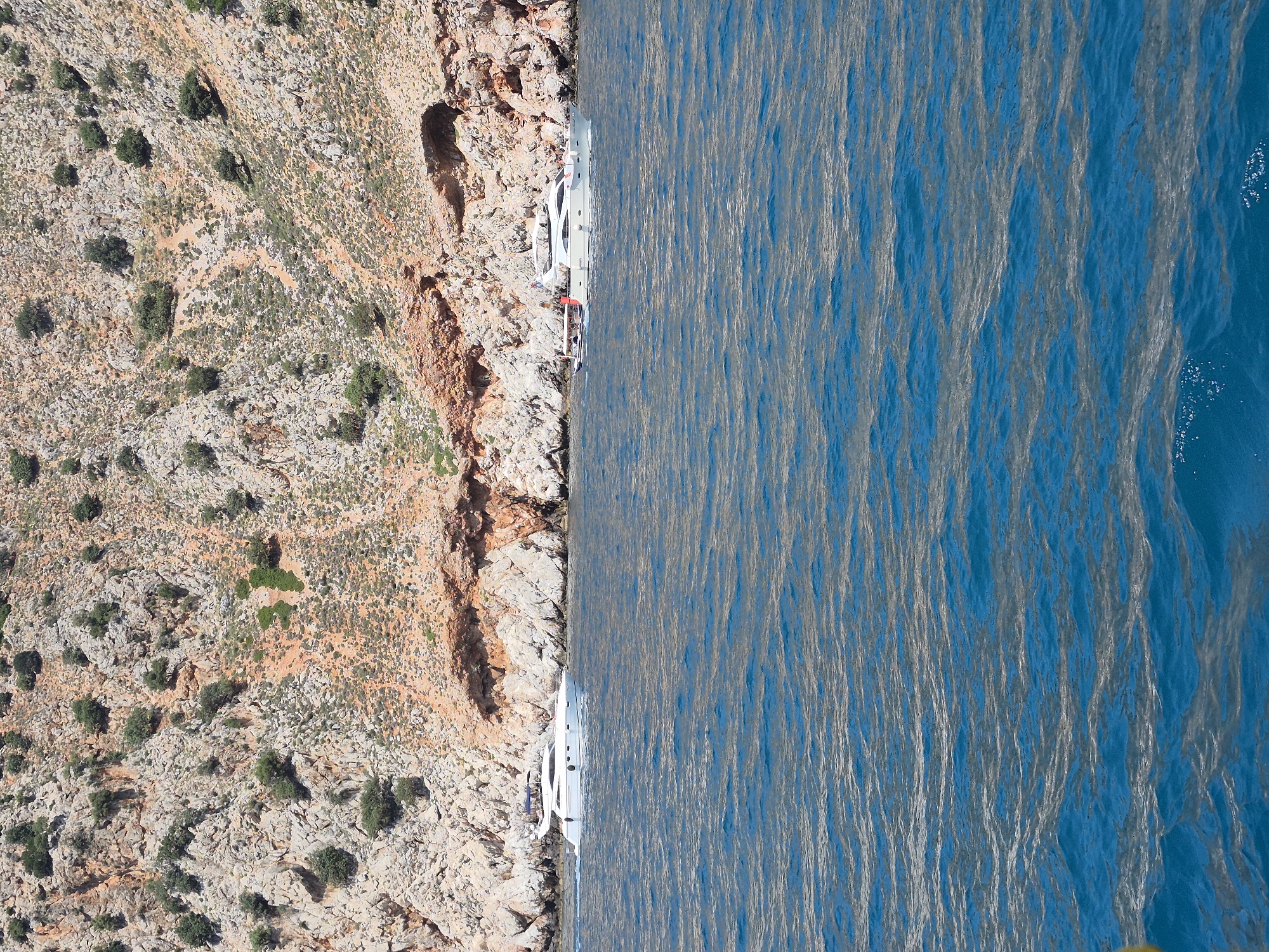 The image is an aerial view, rotated 180 degrees, showing a coastline with a rocky, sparse cliff on the left and the Mediterranean Sea on the right.
Along the edge where the cliff meets the sea, several white yachts are docked.
The cliffside is composed of light brown and gray rock interspersed with some green vegetation. A small cave or crevice is visible in the cliff face.
The sea is a deep blue color, rippled with light reflections from the sun.
The image suggests a location in a coastal region, possibly within the city of Alanya, Turkey, given the characteristic landscape and presence of yachts typical for tourist locations with access to the Mediterranean Sea. There are no visible people, and the overall impression is of a summer tourist spot.