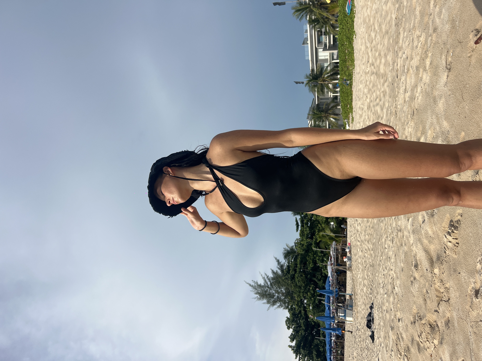 A woman is on a sandy beach in Choeng Thale, Thailand. She is wearing a black swimsuit and a black hat. She is standing with one hand raised to her face. Behind her are trees and buildings. The sky is blue. Beach umbrellas and lounge chairs can be seen nearby.