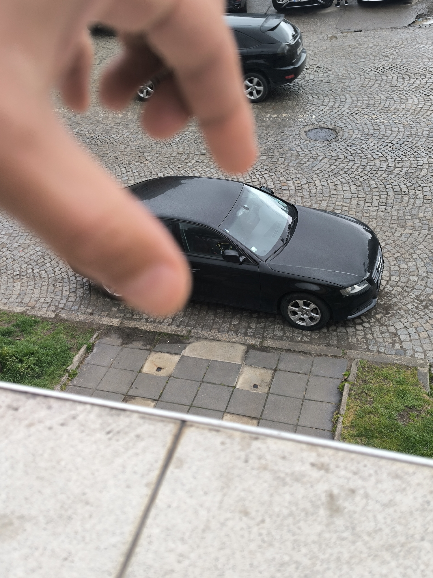 The photograph, taken from an elevated position, depicts a street scene in Sofia, Bulgaria. The foreground is dominated by a blurry human hand extending into the frame. Below and beyond the hand, a black Audi A4 is parked on a cobblestone street. Next to the car, there's a sidewalk with grass patches nearby. In the background, partially visible are parts of an SUV, and other cars further back in the scene. The weather appears overcast, casting a muted tone over the cobblestone street and the surrounding buildings. A manhole cover is visible on the street near the SUV.
