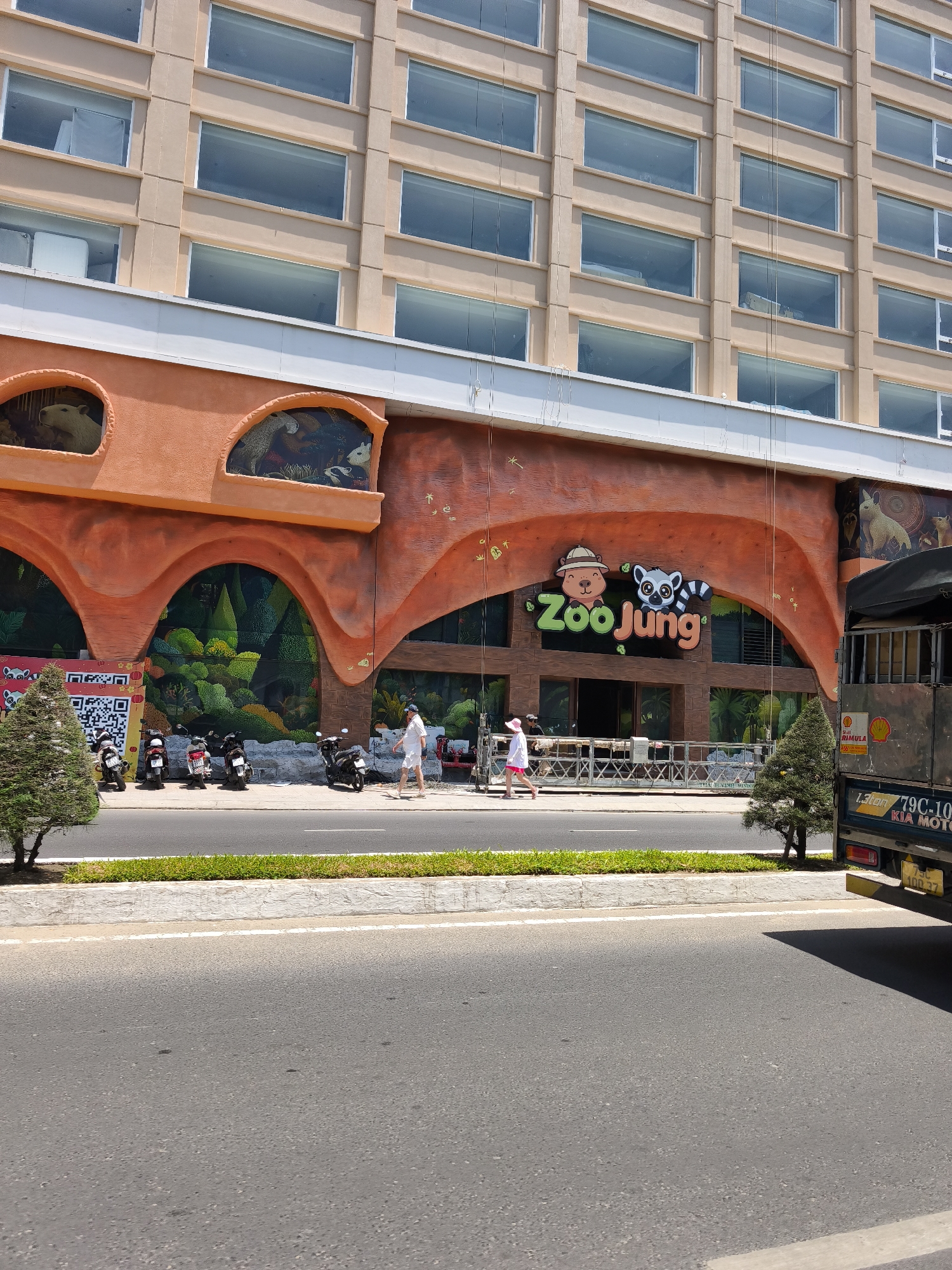 A slightly low-angle, medium shot captures the front of the "Zoo Jung" amusement attraction, which is part of a larger building. The building is a multi-story structure with many horizontal windows on its upper floors, framed by beige-colored panels. The lower portion of the attraction features a vibrant orange facade with arched openings that reveal a mural of a lush green forest. Above these arches, there are smaller arched windows displaying animal figures within them. The "Zoo Jung" logo, featuring cartoon characters and colorful text, is prominently displayed over the entrance area.
In front of the attraction, a paved sidewalk runs along the street. Several motorcycles are parked on the sidewalk. A man in a white shirt and shorts walks along the sidewalk away from the entrance. To the right, a dark-colored truck with a cargo bed is parked on the side of the road. The truck has yellow license plates with Vietnamese characters. The foreground shows a paved road with white lane markings, bordered by a green strip of grass and a white concrete curb. The overall scene suggests a commercial or entertainment area in an urban setting. The location is identified as Bac Nha Trang Ward, Vietnam.