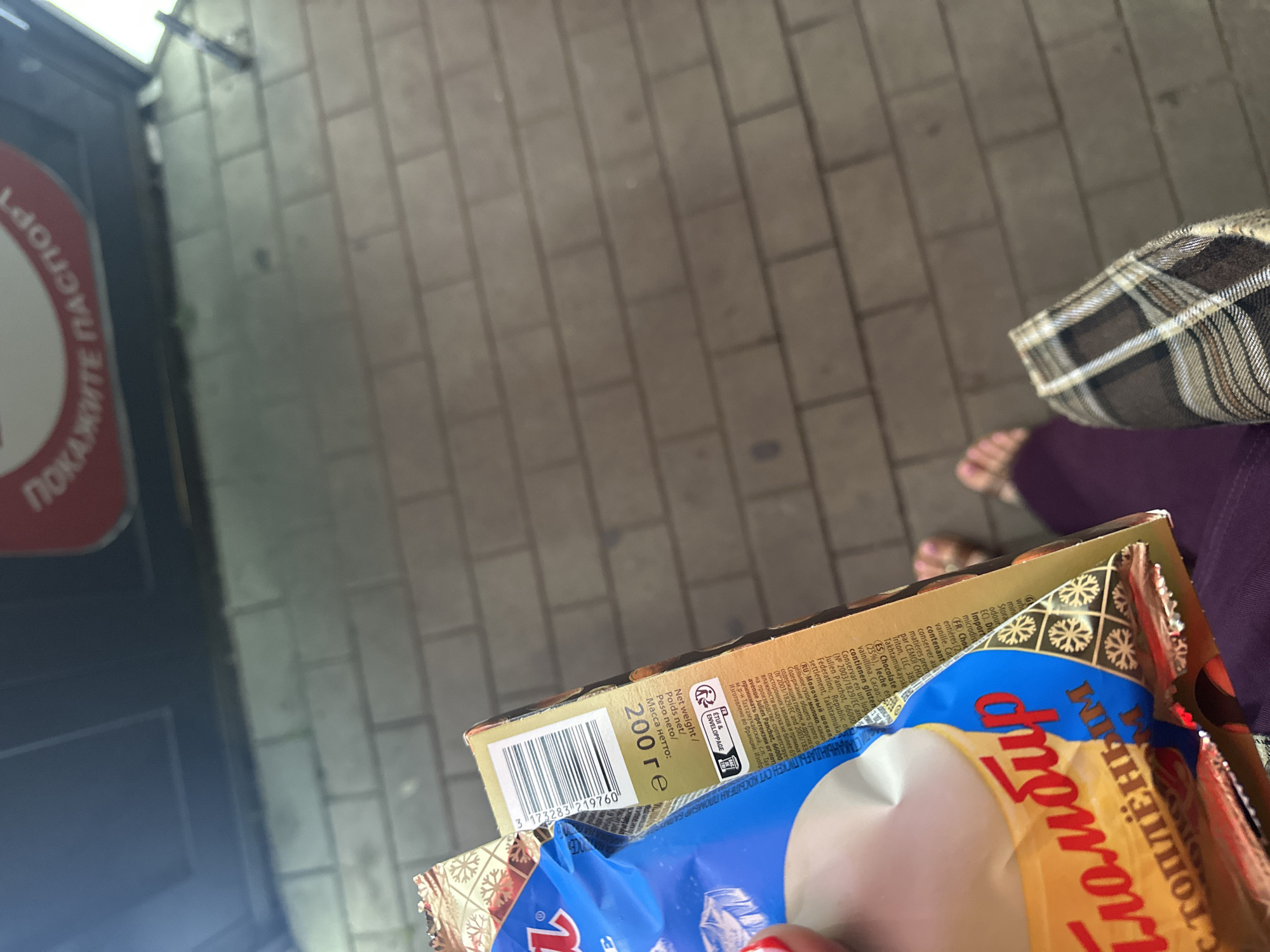The image shows a close-up view of a person holding a packaged food item, possibly ice cream, with Russian text on it. The packaging is yellow and blue and appears to be a brand of ice cream. There is also a barcode visible on the side of the package. In the background, there is a brick wall, suggesting an outdoor setting. The person's legs and feet are visible at the bottom of the frame, indicating they are wearing sandals and purple pants. The overall scene is casual and appears to be taken outdoors on a sidewalk or pathway. There is a sign above the brick wall with Russian text which is blurry but appears to be an advertisement.