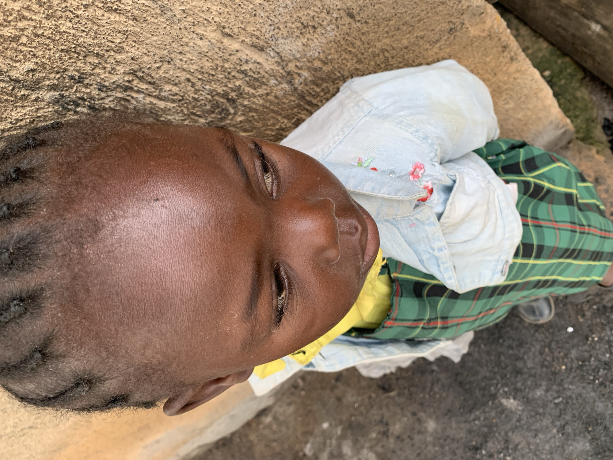 The image shows a close-up, overhead view of a child lying down. The child has dark skin and is wearing a light blue collared shirt with embroidered pink flowers and a green, yellow, and red plaid skirt. The child's head is tilted back, and their eyes are partially closed, suggesting they might be resting or sleeping. The background consists of textured brown stone or concrete on the left and grey gravel on the right. The scene appears to be outdoors, possibly in a simple, rustic environment given the rough textures of the ground. The lighting suggests it could be daytime, but the specific time and weather conditions are not clear. There is no visible text in the image.