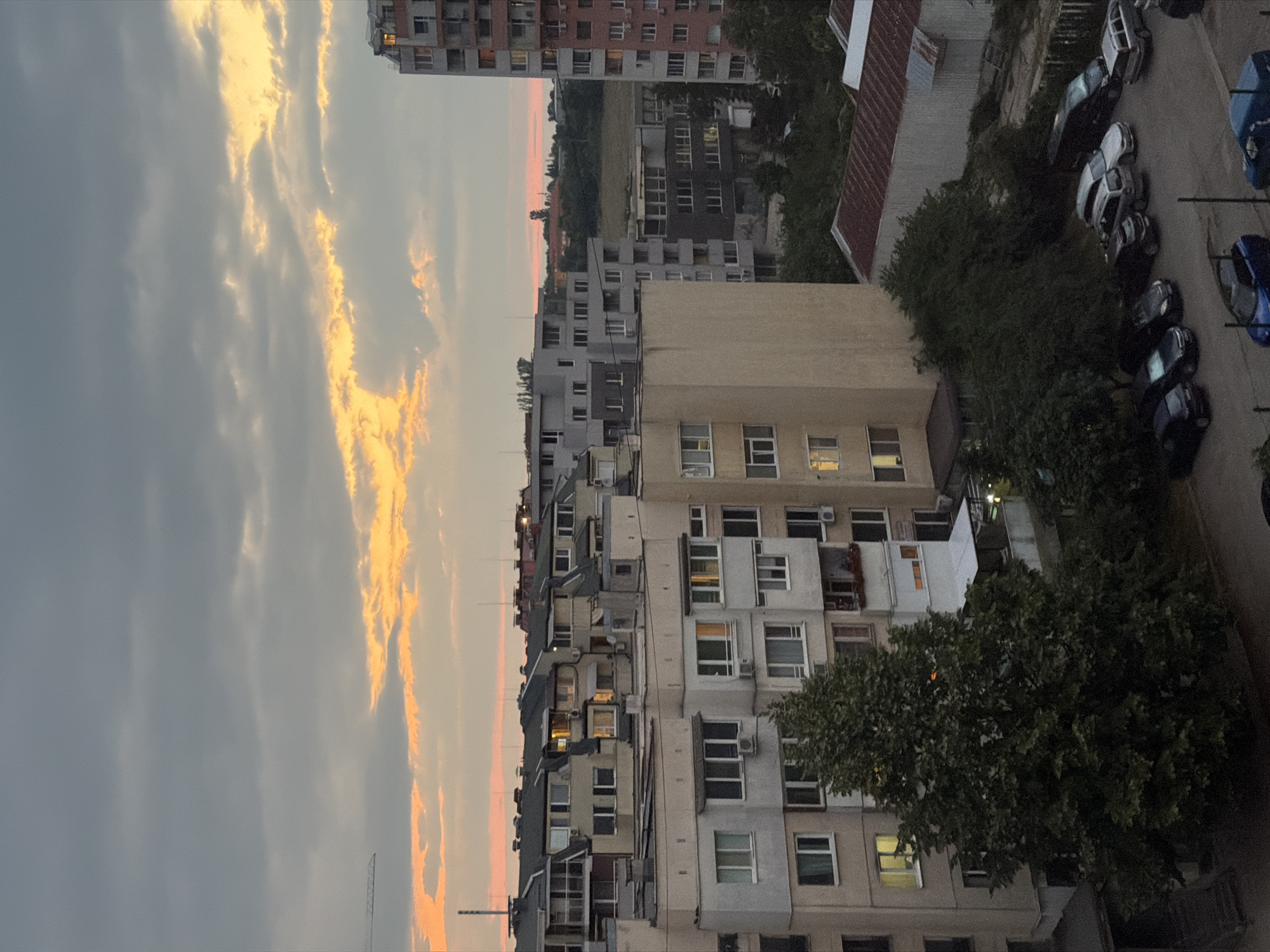 The image depicts an urban scene in Sofia, Bulgaria, during what appears to be sunset or twilight. The sky is dominated by gray clouds with streaks of orange and yellow light breaking through, indicating the setting sun. In the foreground, there are several apartment buildings of varying heights and architectural styles, common in Soviet-era or post-Soviet construction. These buildings are densely packed, with visible windows, balconies, and air conditioning units. Some windows are illuminated, suggesting indoor activity.
The buildings are varied in color, featuring shades of beige, gray, and pink. One tall, modern pink building stands out on the right side of the frame. Below the buildings, a tree with dense foliage occupies a significant portion of the lower left side. In the lower right section, a parking lot with several cars is visible. The cars are of various makes and models, parked in an organized manner. Beyond the immediate buildings, a field or undeveloped land is visible, with some distant structures and vegetation. A construction crane is partially visible in the upper left corner, suggesting ongoing development in the area. The overall impression is of a residential district in a European city, characterized by its mix of older and newer apartment blocks and the encroaching dusk.