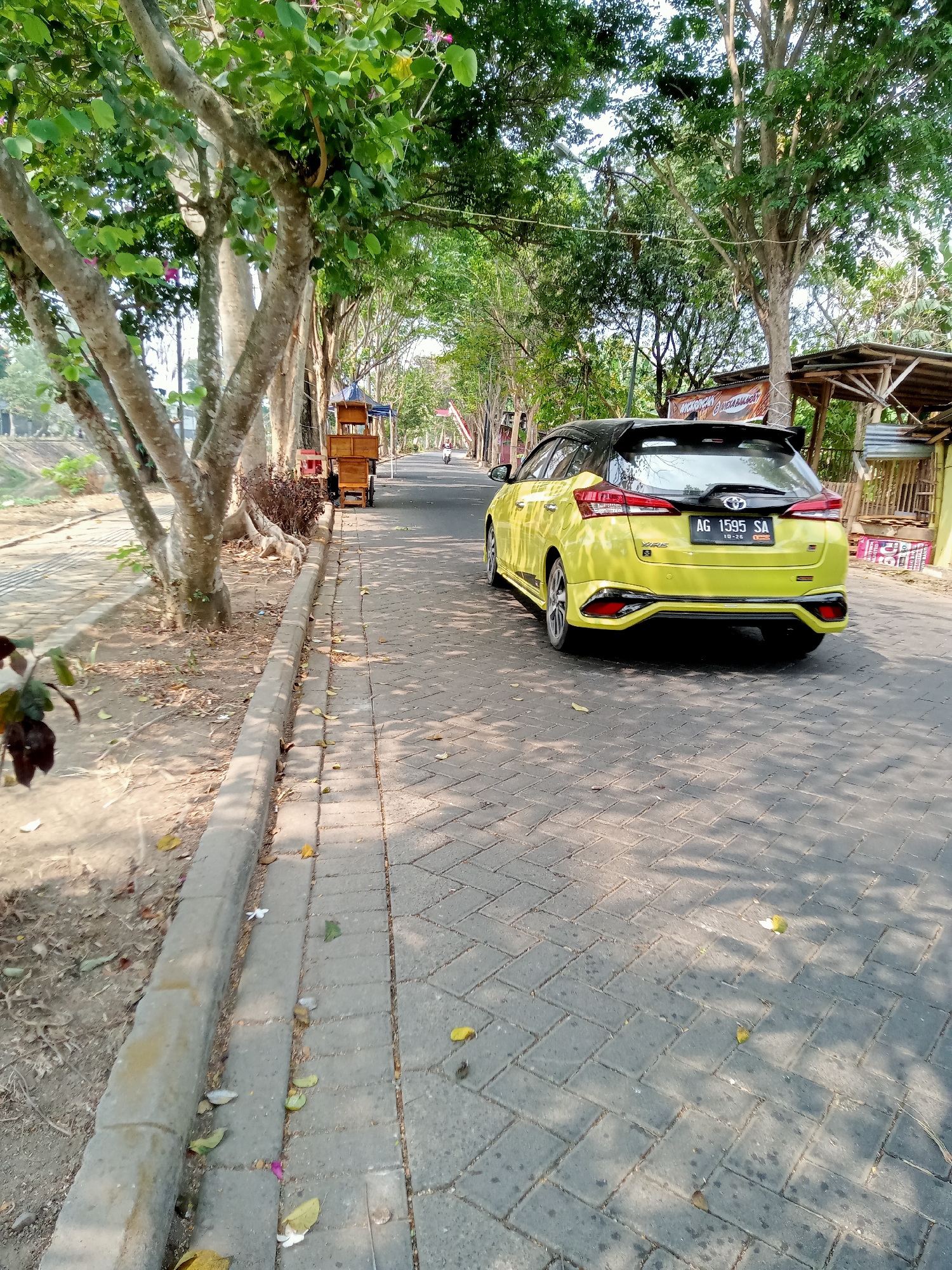 A bright yellow Toyota Yaris car is parked on a paved road lined with trees. The car has a black spoiler and black accents on its body. The license plate reads "AG 1595 SA". To the left of the road, there is a sidewalk with a curb, beyond which is a dirt area with trees. In the distance, there are some wooden carts and stalls. The scene appears to be an outdoor public area, possibly a park or a roadside market, in Tulungagung, Indonesia, given the license plate format. The lighting suggests it is daytime, with sunlight casting shadows from the trees on the road.