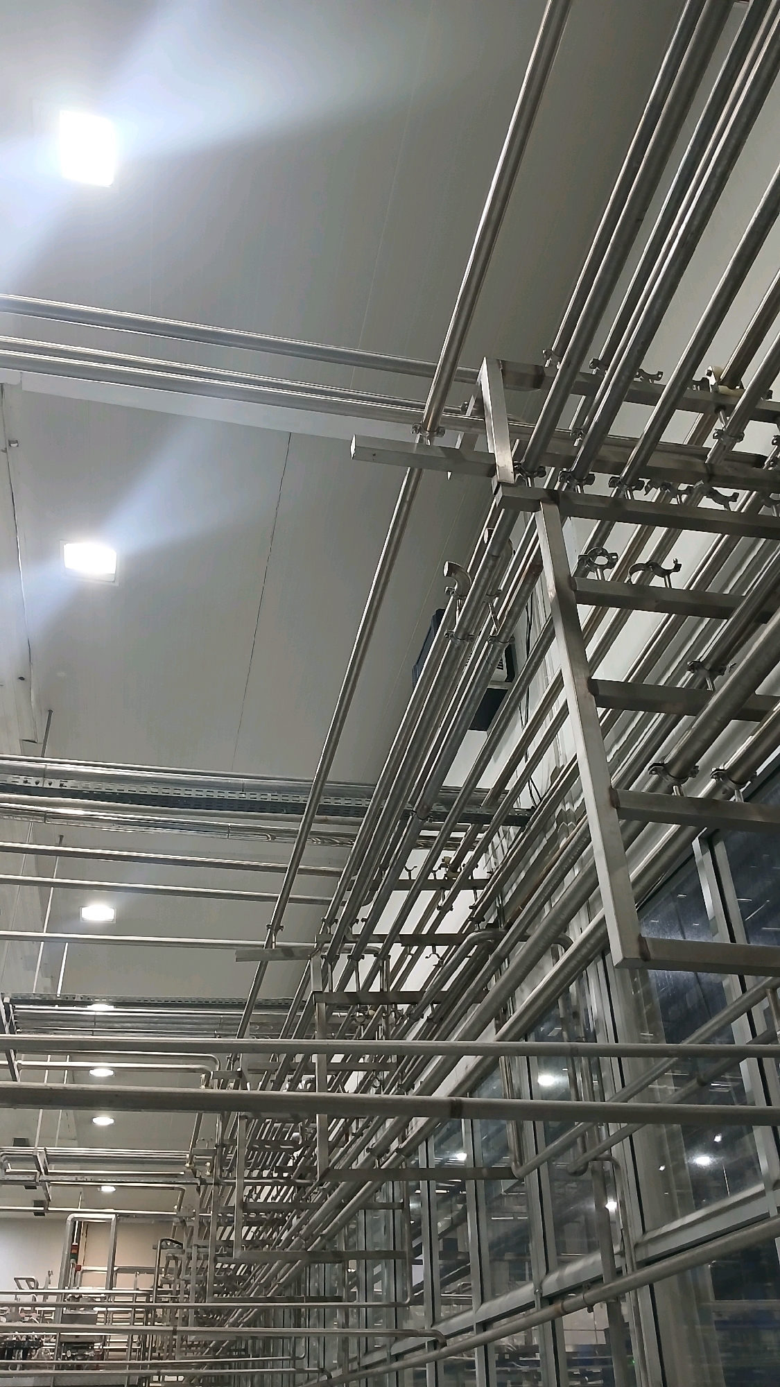 The image depicts an industrial interior, likely a factory or processing plant, characterized by an extensive network of stainless steel pipes suspended from the ceiling and supported by metal structures. The lighting appears to be artificial, with bright rectangular ceiling fixtures casting a cool light. The environment is clean and functional, with a focus on infrastructure rather than decoration. In the lower left of the image, some machinery and equipment are visible, suggesting an operational facility. The overall impression is one of order and efficiency, typical of a manufacturing or production setting. The location is identified as Gadobangkong, Indonesia.