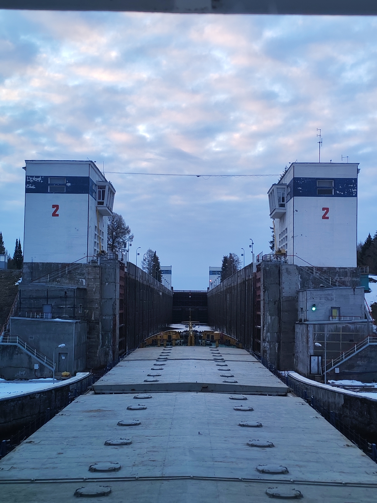 This image depicts a large vessel, likely a cargo barge or ship, positioned within a canal lock in Ankhimovo, Russia. The scene is framed by two tall, white control towers, one on each side of the lock. Both towers feature a prominent horizontal blue stripe and a large red letter "Z" painted on their white facades. Each tower also has an enclosed observation or control cabin extending from its upper section. A thin cable or wire spans horizontally between the tops of the two towers.
The lock walls are constructed from weathered concrete and dark metal, with snow visible on the surrounding ground and some ledges, indicating cold weather conditions, likely winter or early spring. The sky above is overcast with soft, diffused light, suggesting either early morning, late afternoon, or simply a heavily clouded day.
The deck of the vessel, which appears to be flat, is visible in the foreground, featuring multiple oval-shaped hatches. On the right side, a small green light is visible on one of the auxiliary buildings at the base of the control tower. Steps and various structures are integrated into the concrete banks of the lock. Beyond the immediate infrastructure, evergreen trees and some bare deciduous trees dot the landscape. The overall setting portrays robust industrial waterway infrastructure in a cold, functional environment.