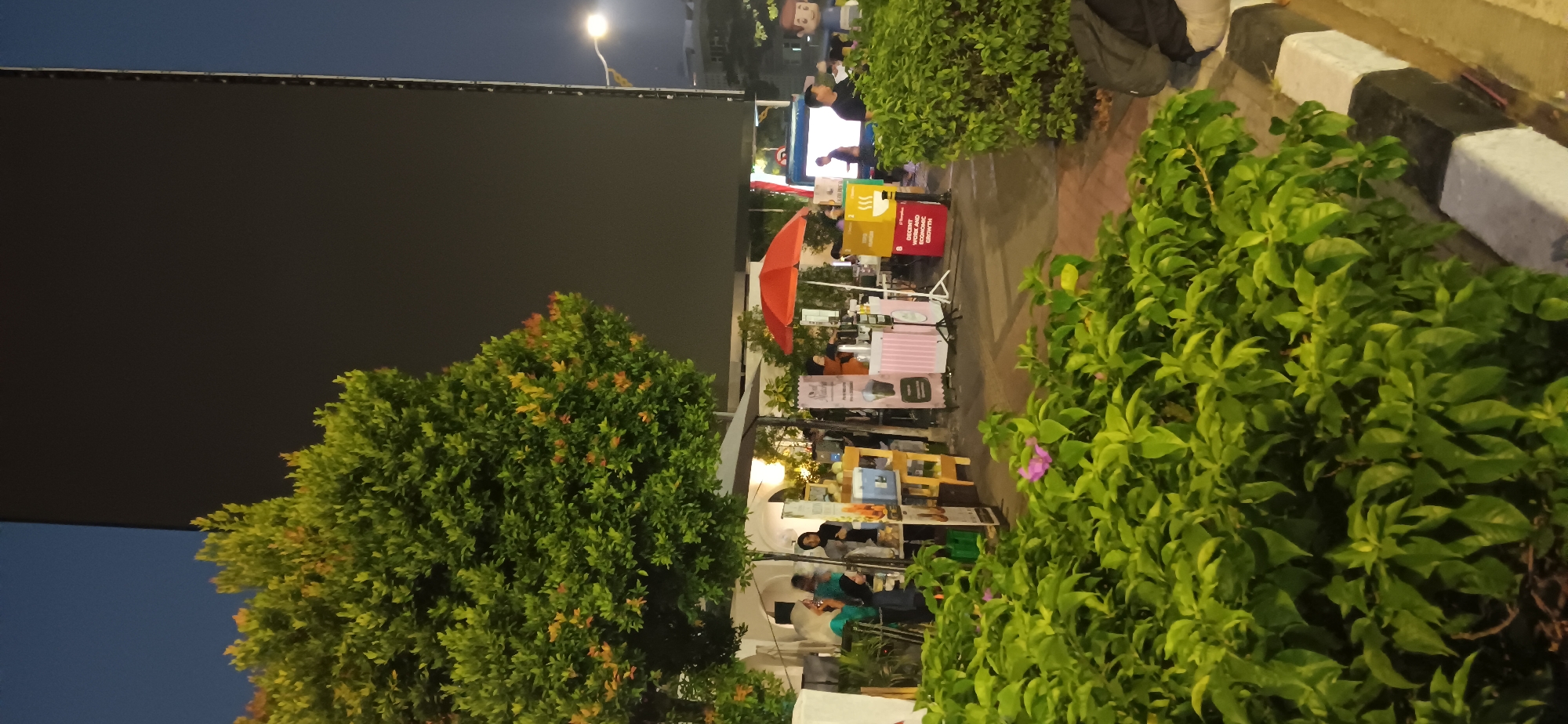 This is an exterior, nighttime photograph taken in South Jakarta, Indonesia, likely at a market or event. In the foreground, lush green foliage with a small pink flower obscures the bottom of the frame. Behind the foliage, there are several stalls set up. One stall has a pink and white striped facade and a red umbrella. Another stall, slightly to the left, is a wooden structure with items displayed on shelves. People are visible at and around these stalls. To the right of the stalls, a large, dark rectangular screen dominates the upper portion of the image, with a street light visible to its right. The background suggests an urban environment with some buildings and streetlights.