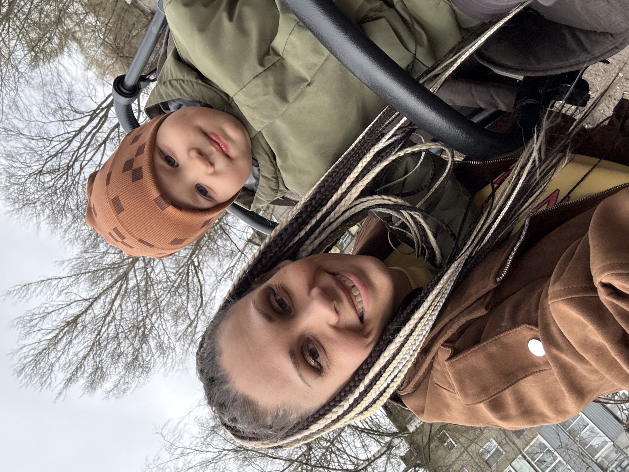 An adult woman and a young child are captured in a selfie-style portrait taken outdoors in Zhukovo, Russia. The image is oriented vertically, with the subjects looking up towards the camera. The woman, positioned on the right, smiles broadly at the viewer. She has long, multi-toned braids in shades of black, brown, and light blonde. She is wearing a brown, suede-like jacket.
The child, a toddler, is on the left, looking towards the camera with a calm expression. The child wears a sage green winter jacket and a light brown beanie adorned with a subtle geometric pattern. A black, curved bar, possibly part of a stroller handle or a play structure, is visible in the foreground, extending from the left to the right side of the image, positioned between the two individuals.
The background reveals a light grey, overcast sky, characteristic of winter or early spring. Bare tree branches, devoid of leaves, are prominent against the sky, suggesting a cool climate and time of year. In the lower background, residential buildings with multiple windows are faintly visible, indicating an urban or suburban setting. The scene conveys a casual, pleasant moment between the two individuals. There is no visible text.