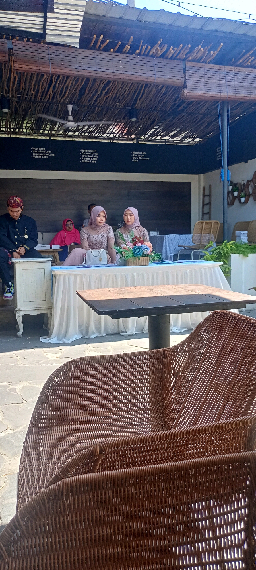 In Desa Ponokawan, Indonesia, an outdoor gathering is underway. A man in traditional attire sits on the left, while two women in headscarves are seated behind a long table covered with a white tablecloth. A floral arrangement sits on the table between them. A person in a pink headscarf is visible in the background. The setting appears to be a cafe or restaurant with a roof made of bamboo and straw. Wicker chairs are in the foreground, and a table with a dark top is set up behind them. A menu is visible on a black board behind the seated individuals.