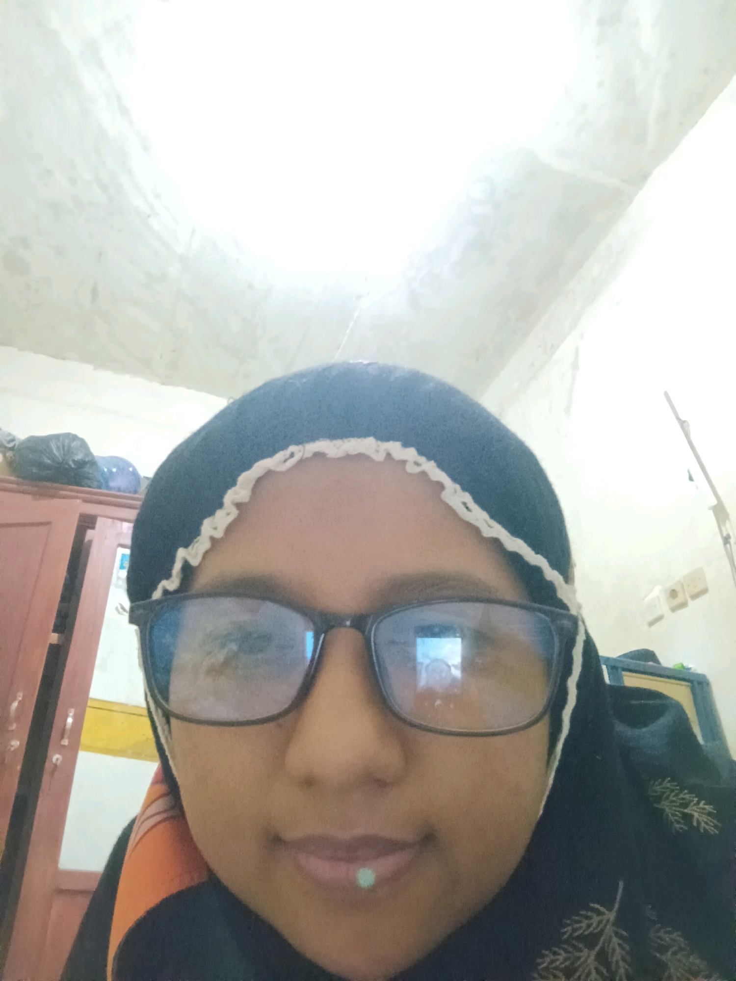 A close-up selfie of a young woman wearing a dark hijab with a white trim and dark-rimmed glasses is taken from a low angle. Her face is centered in the frame, with the ceiling of a room visible in the background. The woman has a small, light-colored stud on her lower lip. Reflected in her glasses is an image of another person. To the left of the woman, a portion of a wooden cabinet or wardrobe is visible. The environment appears to be indoors, with a wall and a light fixture visible in the background. The image does not contain any specific information about the city of Sarangan, Indonesia.