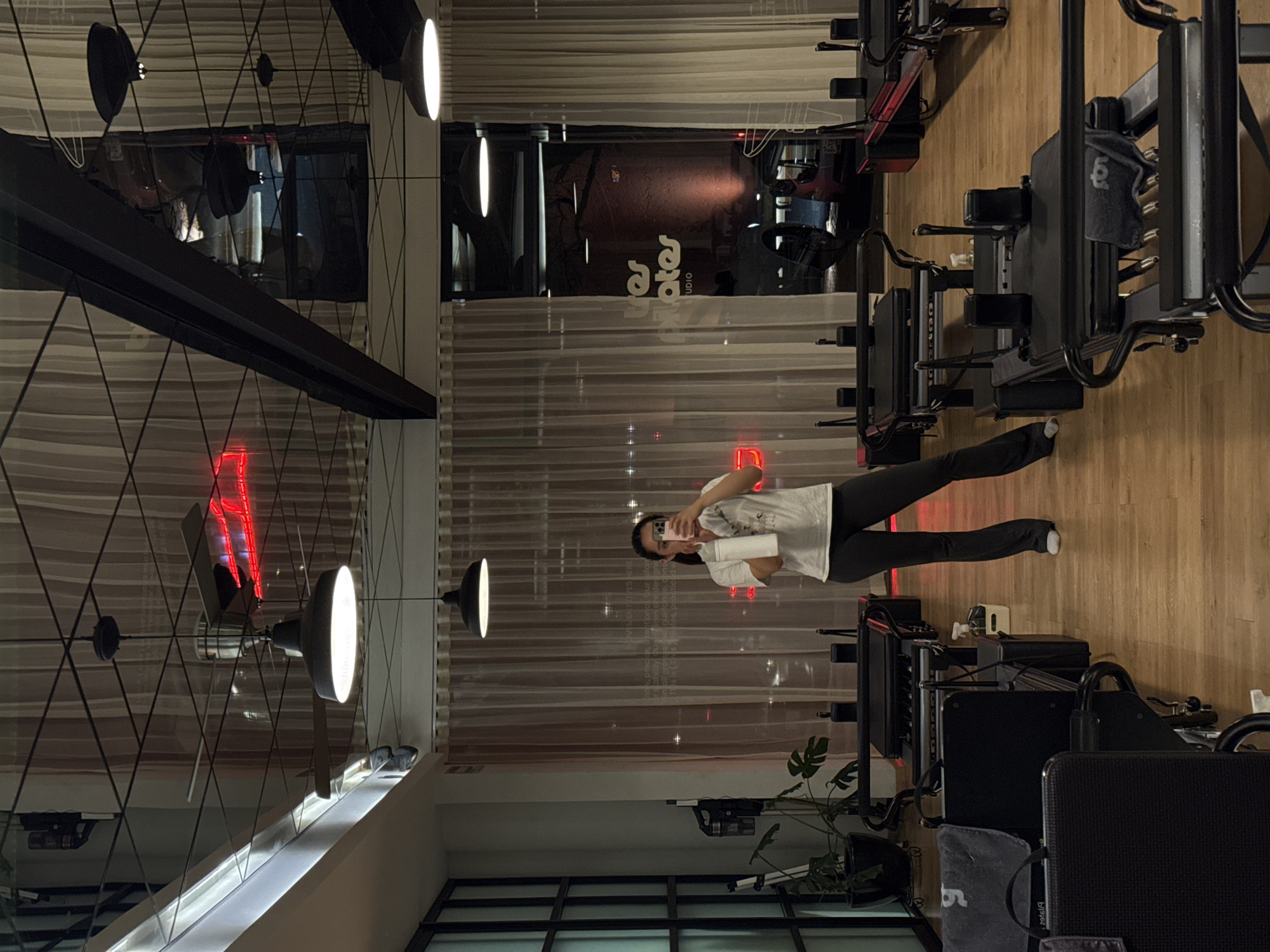 An evening indoor shot in a Pilates studio in Puebla, Mexico, shows a woman taking a mirror selfie in a large window. The woman, wearing a white t-shirt and dark leggings, holds a white water bottle or cup in one hand and her phone to her face with the other. She stands amidst several dark-colored Pilates reformer machines, which are arranged on a light-colored wooden floor.
The studio features a modern industrial design with black overhead structures and several illuminated circular pendant lights. The large window reflects elements of the studio's interior, including the ceiling, lights, and a red neon sign that is also partially visible directly through the window. The reflection on the window clearly shows the text "Pilates Studio" in a stylized font, likely from an external sign or light. Another red neon light, possibly spelling "Pilates," is reflected and directly visible.
The scene is lit by a mix of warm and cool artificial lights, suggesting it is nighttime, with the outdoor environment appearing dark through the window. A small green potted plant is visible on the floor near the bottom left. A grey towel with a partially visible logo or text hangs over one of the reformers.