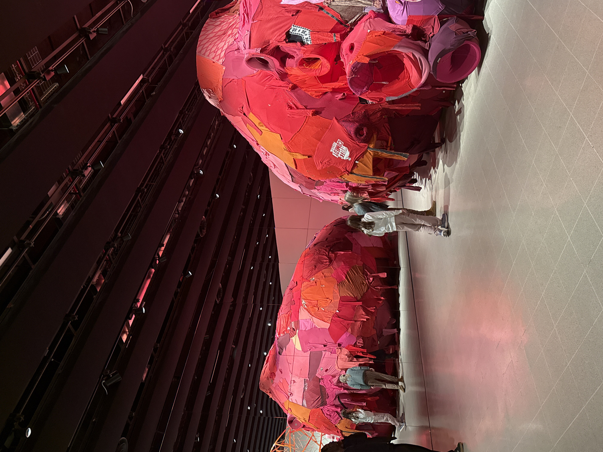 The image captures a section of the Tanks at the Tate Modern, a modern art gallery located in London, United Kingdom. The scene focuses on a large-scale art installation, consisting of two colossal heads crafted from what appears to be recycled or repurposed clothing items, primarily in shades of red, pink, and orange. The heads are suspended from the ceiling and positioned at different heights, creating a dynamic visual impact. Several visitors are observing the artwork. The environment includes exposed architectural elements with a dark, industrial aesthetic, along with modern gallery lighting. The floor has a tiled pattern. The color palette is dominated by the vibrant hues of the artwork and the subdued lighting of the gallery space.
