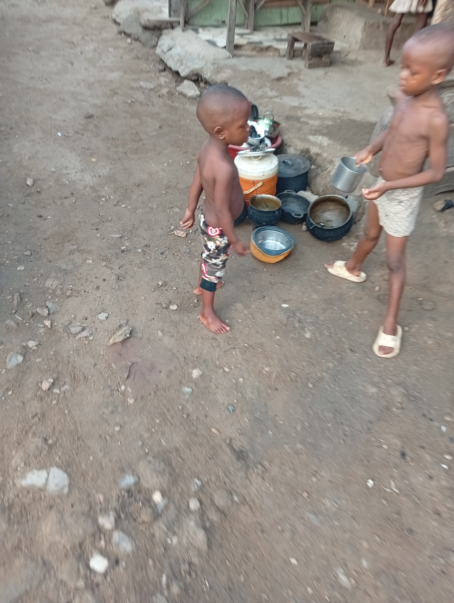 The image captures two young, shirtless boys in an outdoor setting, likely Adankolo, Nigeria. The boy on the left stands barefoot, wearing camouflage-patterned shorts, facing towards the right. The boy on the right wears light-colored, patterned shorts and beige slippers, holding a silver metal cup in his hands, seemingly interacting with the pots on the ground.
They are situated on a dusty, uneven dirt ground scattered with small rocks and debris. In the mid-ground, behind and between the boys, a collection of kitchenware is visible, including several dark, possibly cast-iron cooking pots, a silver bowl set within a yellow plastic basin, and a white and orange plastic container. In the background, hints of wooden structures and another person's legs (possibly an adult) suggest a residential or communal outdoor area.
The soft, ambient lighting indicates it is daytime, possibly early morning or late afternoon. The scene depicts a casual, everyday moment, with the children seemingly engaged in play or a simple chore involving the pots and cups. There is no visible text in the image.