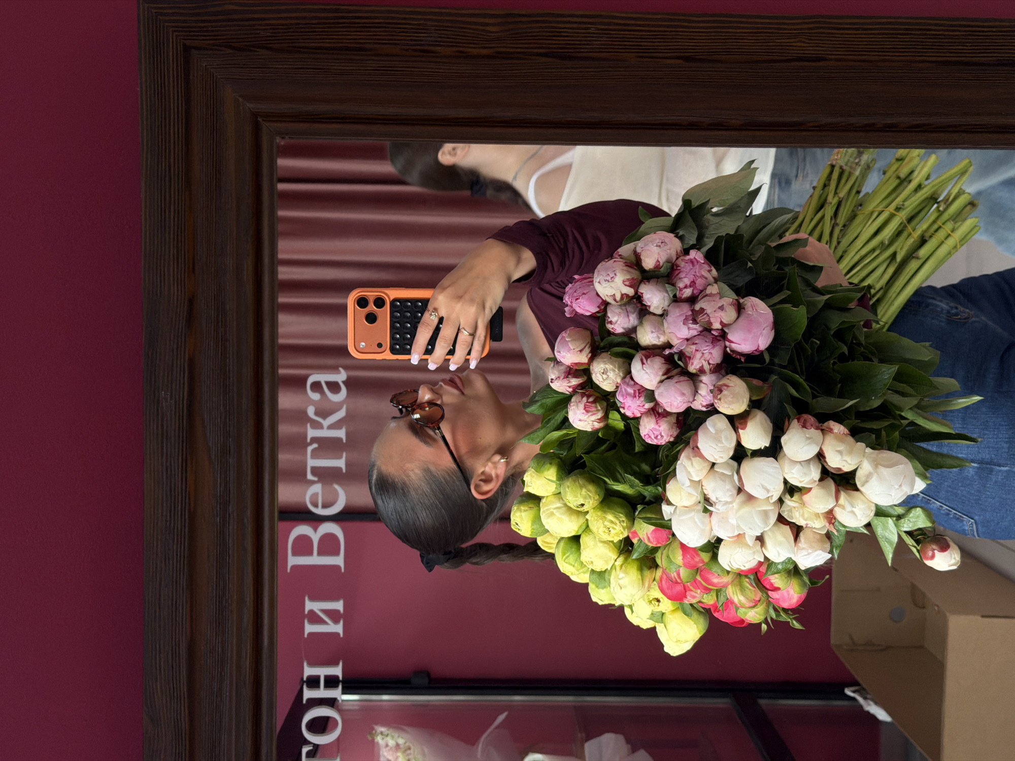 A woman is reflected in a mirror while holding a large bouquet of flowers and taking a photograph with an orange mobile phone. The flowers are predominantly white, pink, and yellow peonies. The woman is wearing sunglasses, a burgundy top, and jeans. The reflection includes a second person wearing a white top and jeans, and a cardboard box. The mirror is framed with dark wood. In the background, painted on the wall, the words "Гон и Ветка" are visible, which are likely the name of a flower shop located in Krasnoyarsk, Russia. The wall is a shade of red.