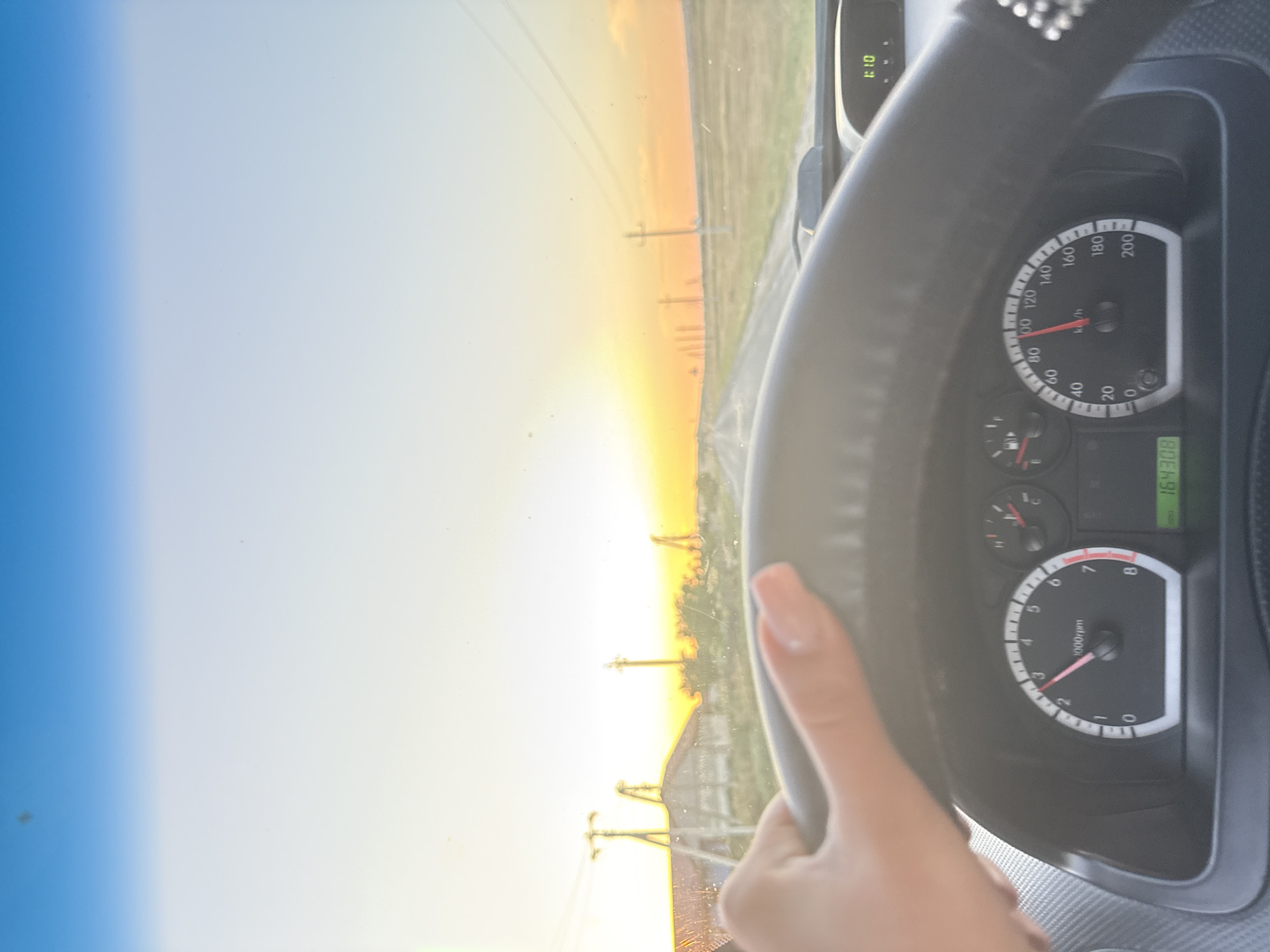 The image shows a first-person view from the driver's seat of a car driving down a road in Cioc-Maidan, Moldova, during sunset. The driver's hand, with manicured fingernails, is visible on the steering wheel. The dashboard displays the car's speed at approximately 100 km/h and the tachometer indicating around 2000 rpm. The digital odometer reads "164308". Outside the car, a rural landscape is visible with a dirt road stretching into the distance, lined with utility poles and fields. The sky is a gradient of blue and orange, indicating the sun is setting on the horizon. There are no other people or vehicles clearly visible in the scene.