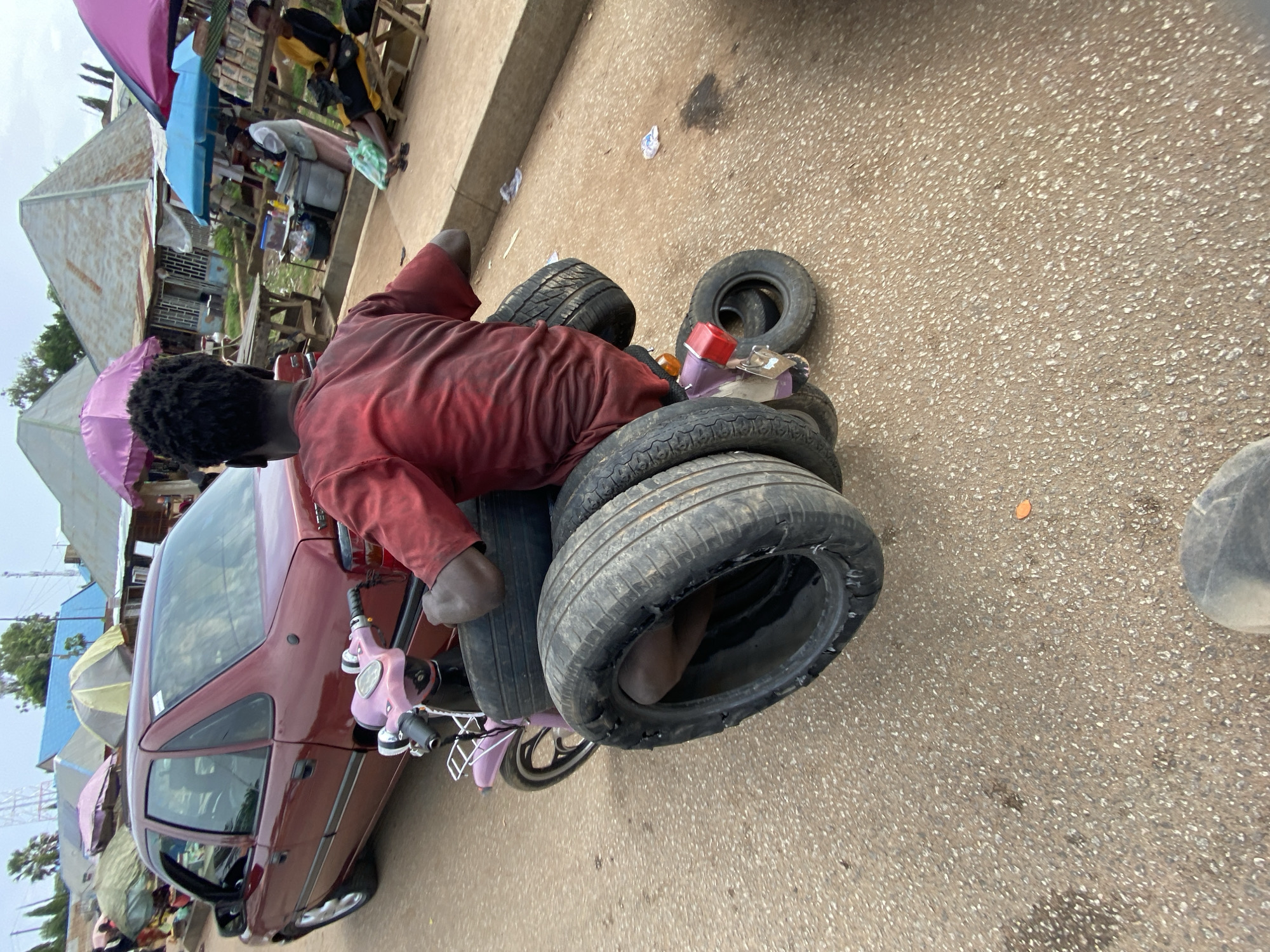 A man in a red shirt sits atop a stack of tires on the ground in Bukuru, Nigeria. He appears to be interacting with a disassembled scooter-like vehicle next to a maroon car. The scene takes place on a paved road near a market with shops and people visible in the background.