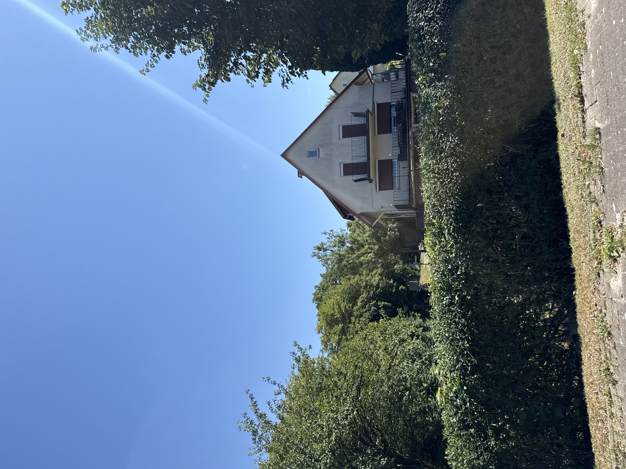 The image captures a bright, sunny day with a clear blue sky and a visible sunbeam entering from the upper right. In the mid-ground, a light-colored, two-story house with a red roof is partially visible, set back from the foreground. The house features balconies with dark railings on both the upper and lower levels, and window shutters are closed on the lower floor. The house is surrounded by lush green trees and a well-maintained hedge in the foreground. The ground in the immediate foreground consists of dry grass and a brick pathway. The scene appears to be located in a residential area.