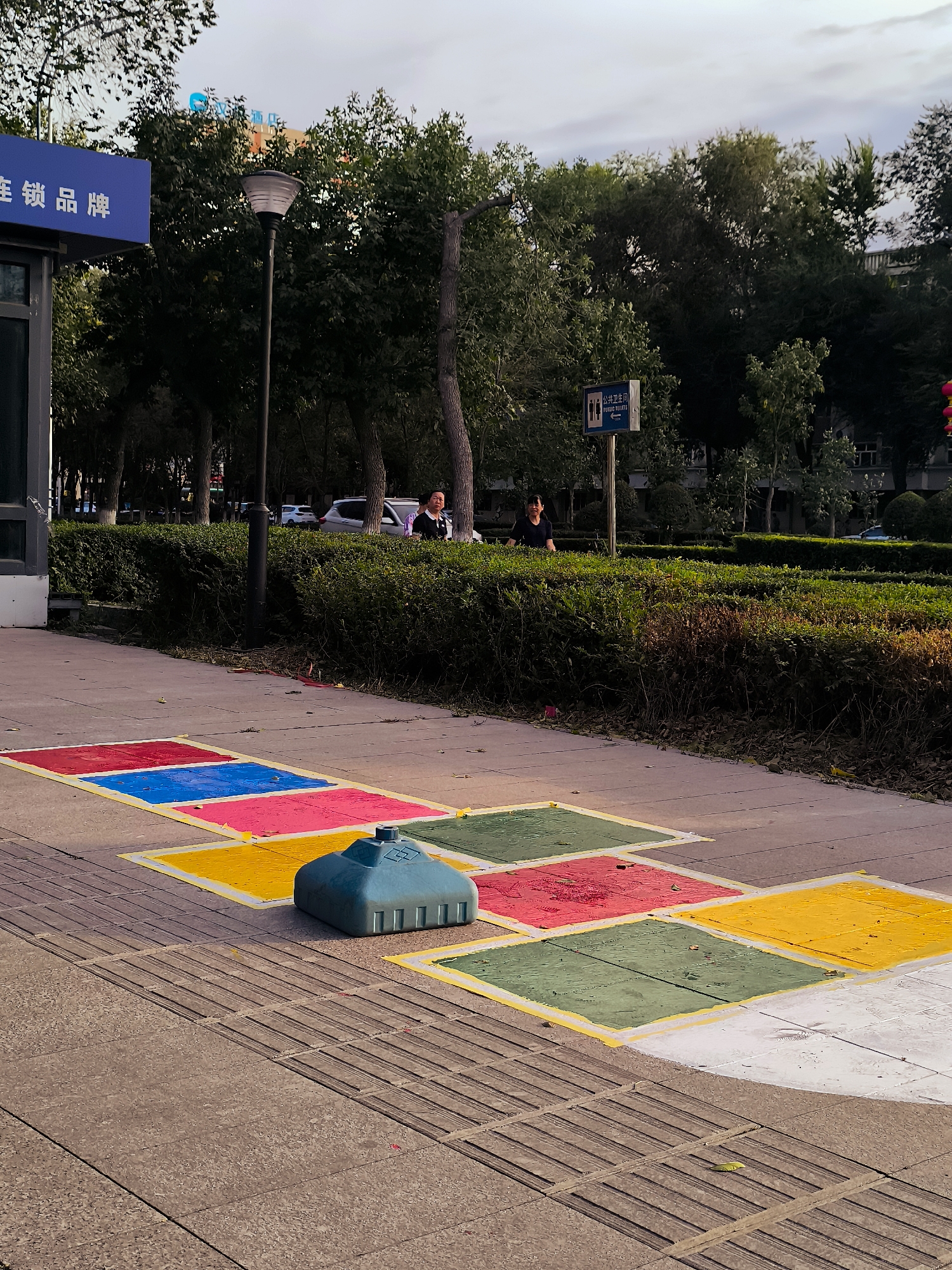 The image depicts an outdoor scene on a paved walkway, possibly a park or a public space, in Ürümqi, China. In the foreground, a hopscotch grid is painted on the pavement with colorful squares of red, blue, yellow, and green, bordered by yellow lines. A turquoise-colored plastic object, possibly a base for a game or playground equipment, sits on the pavement near the hopscotch squares. The walkway is textured with raised linear patterns, indicative of a tactile paving surface, likely for the visually impaired. Beyond the walkway is a neatly trimmed hedge, and behind that, several people are visible. Two women are standing near the hedge, one in a dark top and the other in a lighter top. Further in the background, trees and buildings are visible under a cloudy sky. A lamppost stands near the left edge of the image, and a blue sign with white Chinese characters and an arrow pointing to the left is also present. Another sign, a blue rectangle with white pictograms and text, indicating "Public Toilets," is visible further to the right. Cars are parked or driving in the background, suggesting a roadside or street-adjacent location. The lighting indicates it is either early morning or late afternoon.