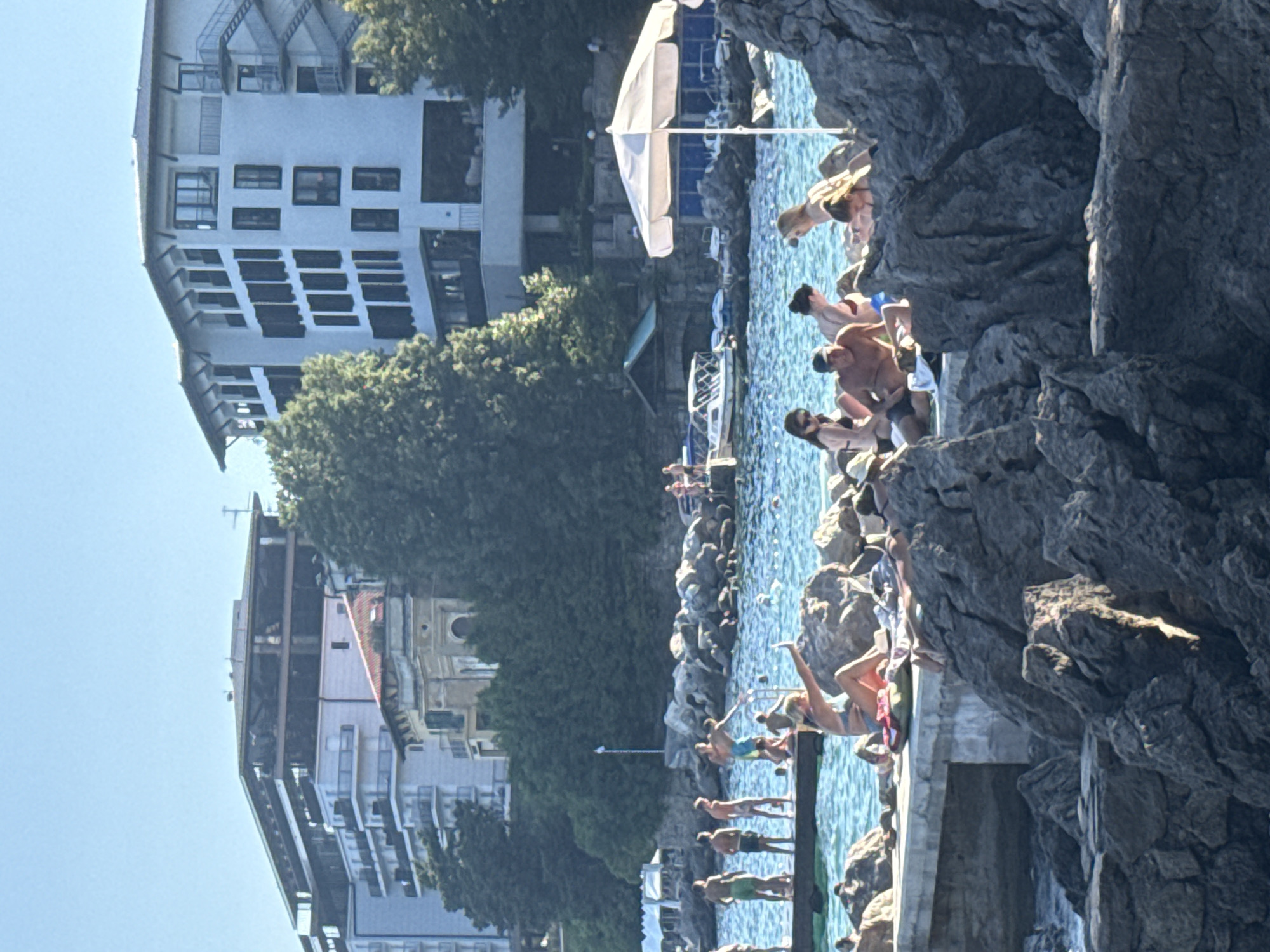 This image captures a scene at a rocky coastline in Grad Opatija, Croatia, bathed in bright, clear sunlight. The water is a vibrant turquoise, with small waves lapping against the shore and a pier jutting out into the sea. Several people are scattered across the scene, engaged in various beach activities. On the pier, a group of men are standing and interacting, with some appearing to be posing for a photo. In the foreground, a woman is sitting on a rock, holding up her arm to take a selfie. Other individuals are lounging on rocks and a concrete platform, sunbathing and relaxing. Behind the beachgoers, a white umbrella shades a seating area near the water's edge. Further back, several buildings are visible, including a tall, modern hotel with balconies and windows, and other apartment-style buildings nestled among lush green trees. A few boats are docked in the water. The overall atmosphere is one of a lively summer day at the seaside.