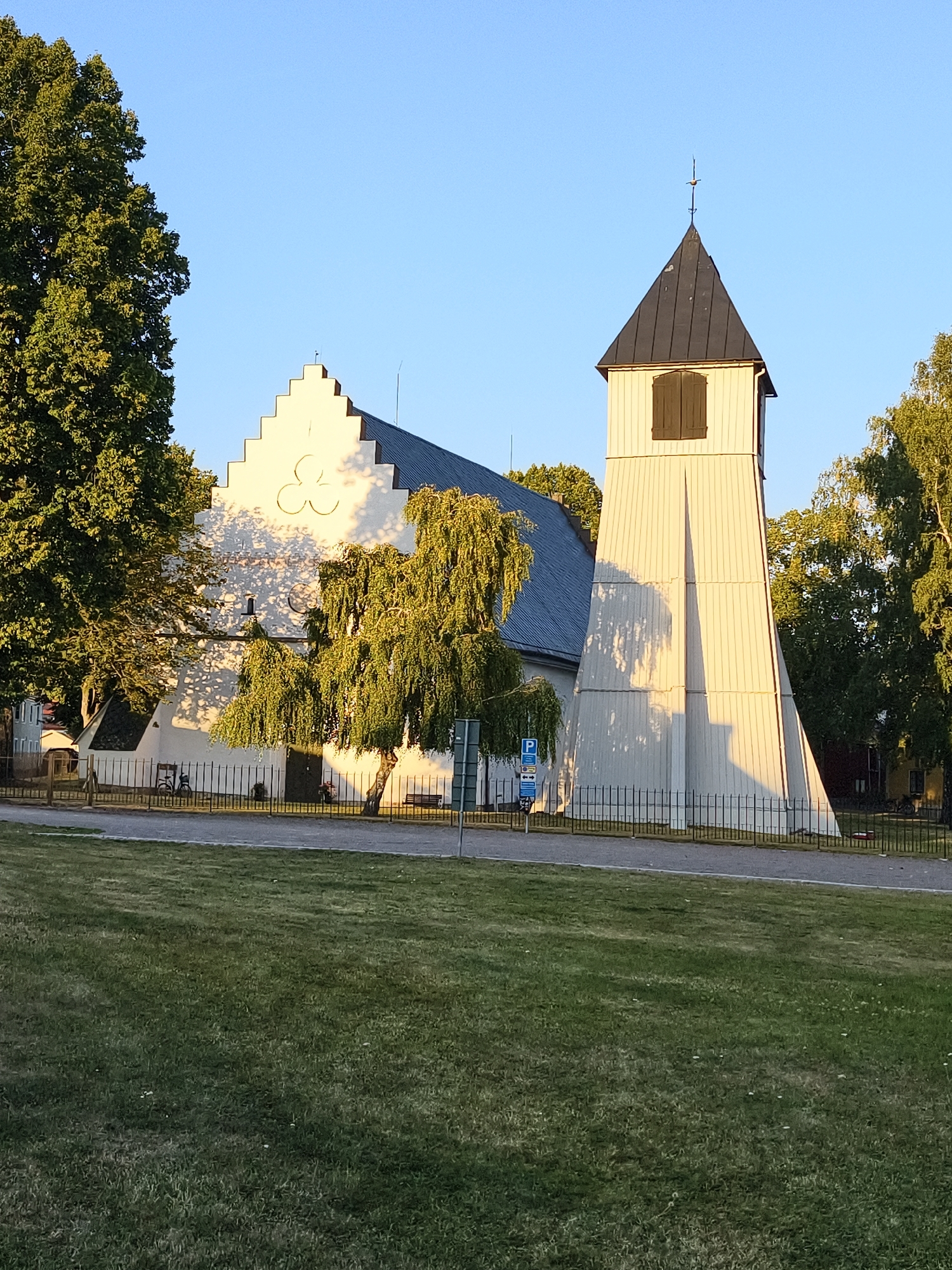 The image shows the church of Östra Husby in Söderköping, Sweden, on a clear, sunny day. The church is primarily white with a dark, stepped gable roof and a tall, white wooden bell tower with a dark pyramidal roof. The sun casts distinct shadows from the trees onto the church building and bell tower. A mature tree with green foliage stands in front of the church, partially obscuring it. A metal fence separates the church grounds from a grassy lawn in the foreground. A blue parking sign is visible near the fence. The overall impression is one of a peaceful, well-maintained historical building in a natural setting.