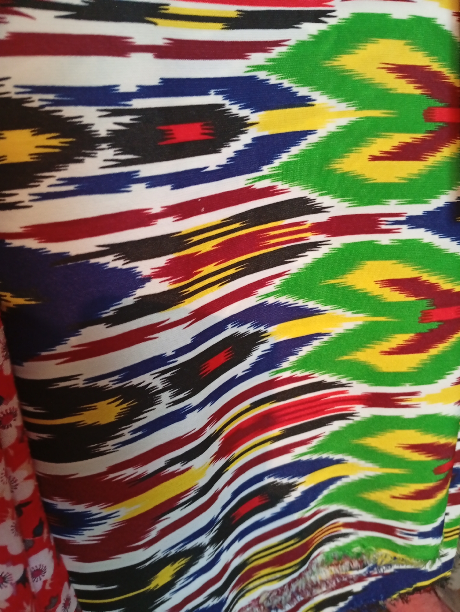 The image shows a close-up of a colorful, patterned fabric. The pattern appears to be a traditional ikat or suzani style, with bold geometric shapes and wavy lines in vibrant colors like green, yellow, red, blue, black, and white. The fabric is draped, creating folds and shadows that highlight its texture and the way the light falls on it. On the left side of the image, a sliver of another fabric with a red background and a white floral pattern is visible. There is no discernible location in the city of Seýdi, Turkmenistan, as the image is focused solely on the textile.