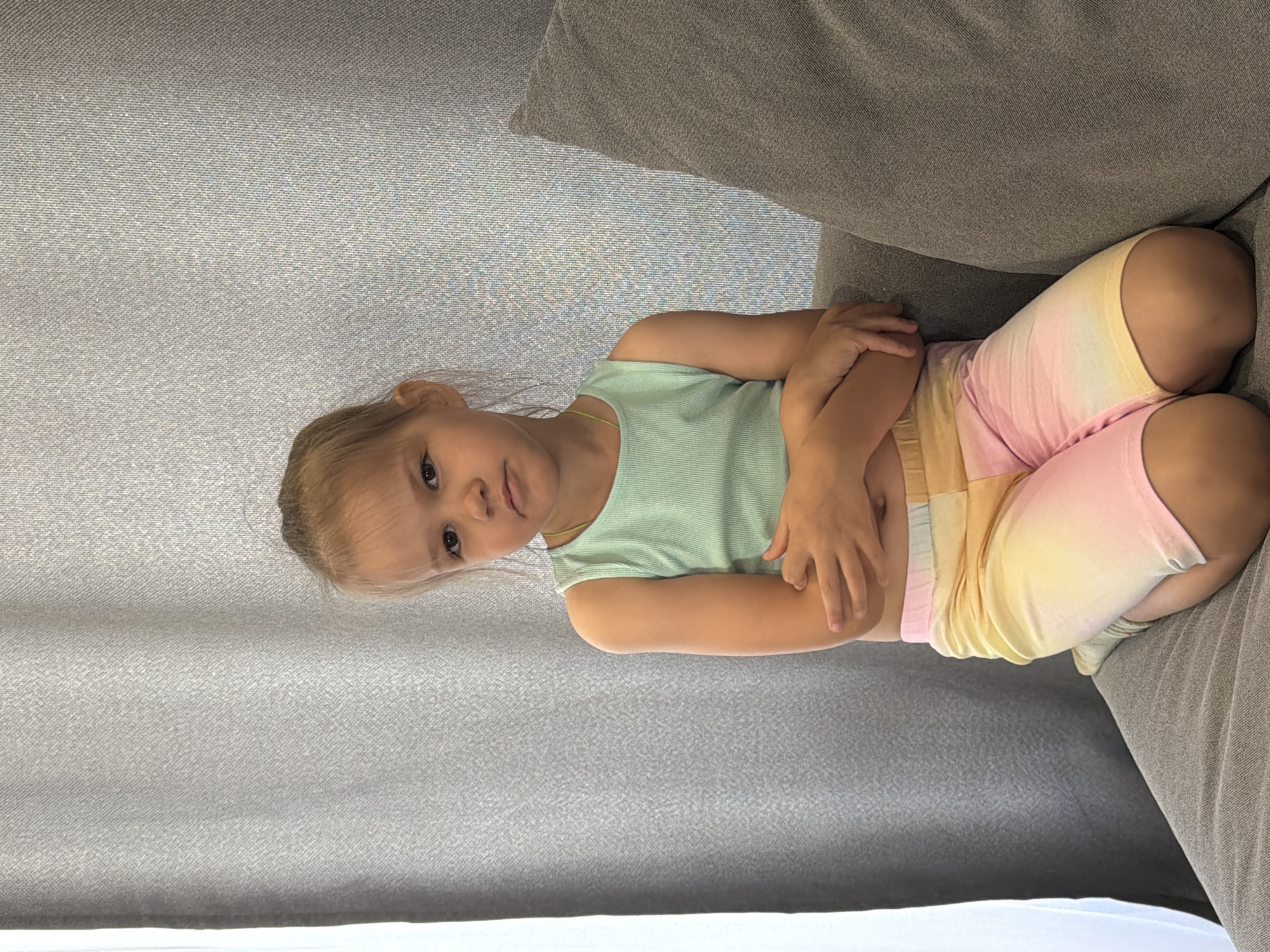 A young girl is resting on a gray fabric sofa. She has fair skin and blonde hair pulled back. She's wearing a light green sleeveless top and pastel tie-dye shorts. Her arms are folded, with her hands resting on her arms. The setting appears to be indoors. There is no information to confirm the location in the city of Chaikovsky, Russia.