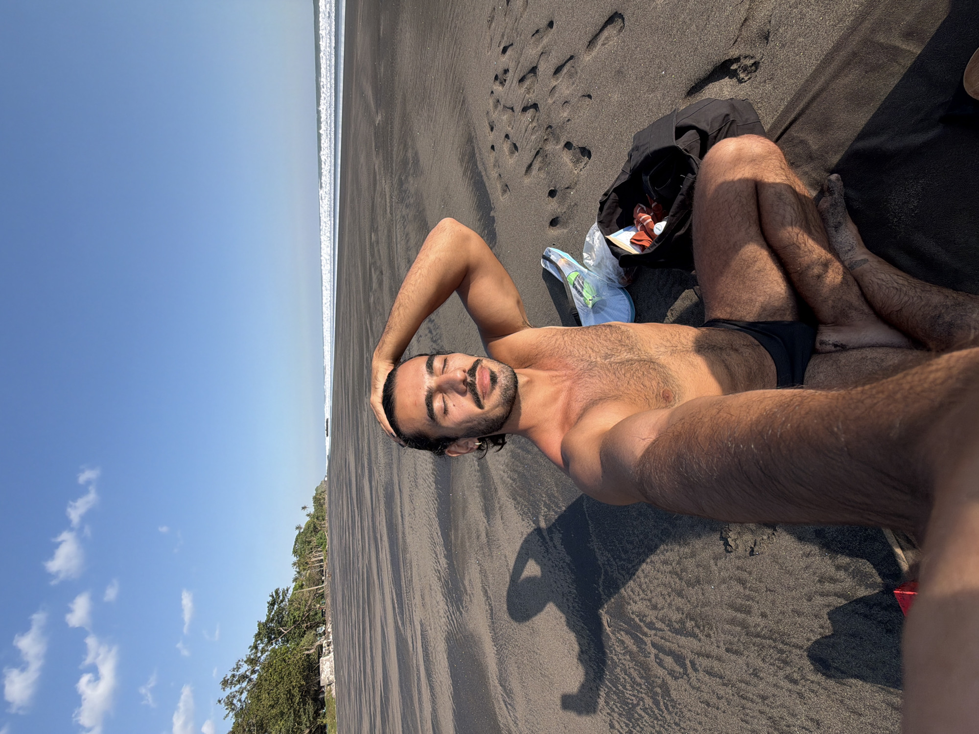 A male individual is depicted in a full-frame selfie, seated cross-legged on a black sand beach in Selemadeg Timur, Indonesia, under a clear blue sky with scattered white cumulus clouds. The man, who appears to be of Mediterranean or Middle Eastern descent, has dark hair, a beard, and a mustache. He is shirtless, wearing dark swim trunks, and has a small silver hoop earring in his left ear. His eyes are closed, and he exhibits a slight smile, with his right hand (viewer's left) resting on his head and his left arm (viewer's right) extended forward holding the camera. A distinct shadow of the man, including his arm and hand holding the camera, is cast on the dark sand to his left. To the man's right, a black open bag contains orange-and-white striped fabric and other items, next to a clear plastic bag holding a white object and a green item. The wide black sand beach extends into the distance, meeting the ocean where white waves break on the horizon. Lush green trees and palm trees line the distant left side of the coastline. The scene is brightly illuminated by direct sunlight.