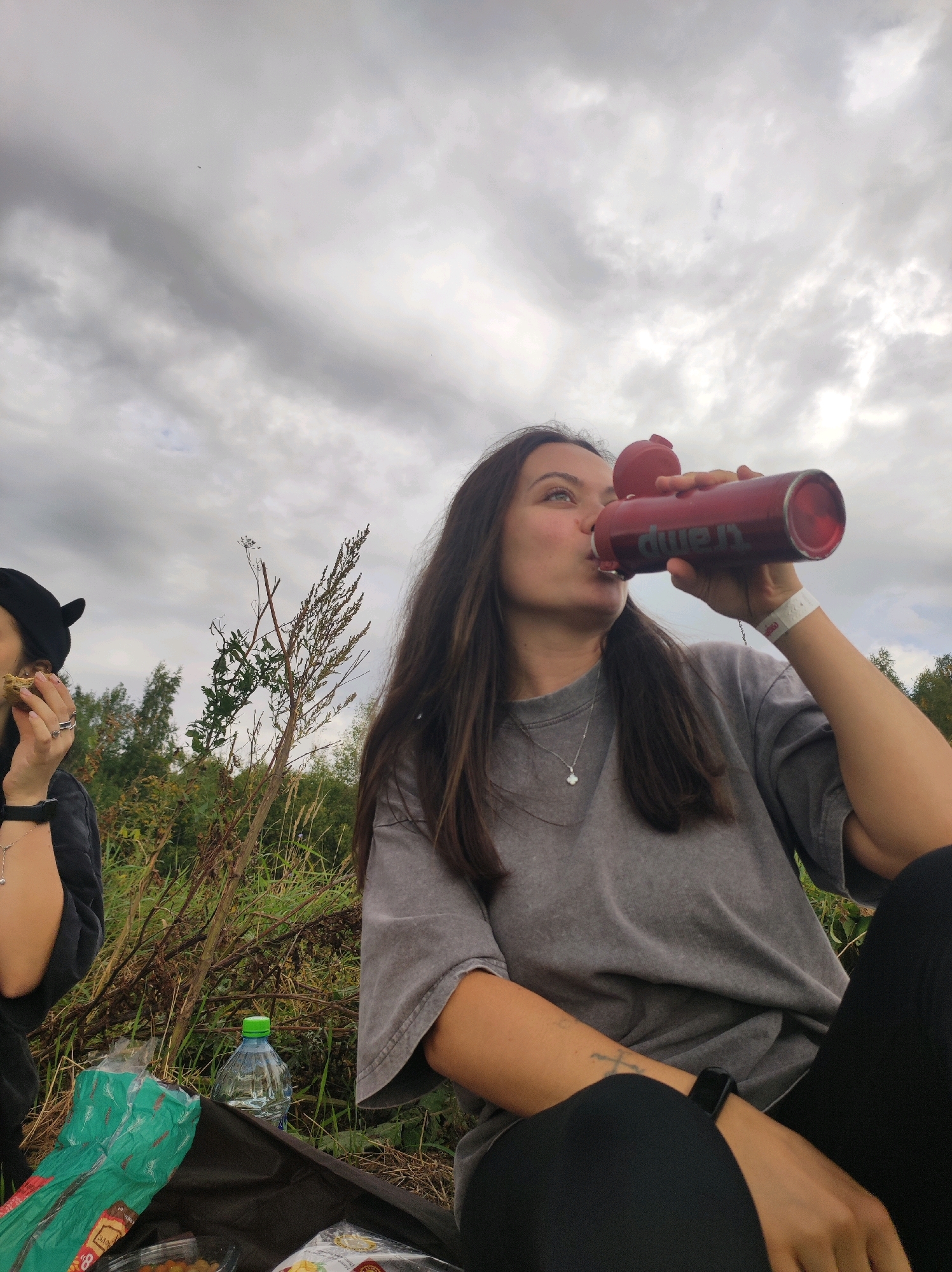 In Italmas, Russia, under a cloudy sky, two individuals are engaged in an outdoor activity, likely a picnic. The foreground is dominated by a woman in a grey sweatshirt, actively drinking from a red water bottle. Beside her, partially visible, another person wears a black cap with ear-like protrusions and is shown consuming food. The environment suggests a natural setting with tall grasses and trees in the background. A black blanket lies on the ground, partially covered by food packaging and a plastic water bottle. The scene is captured from a medium-low angle, focusing on the two individuals and their immediate surroundings.
