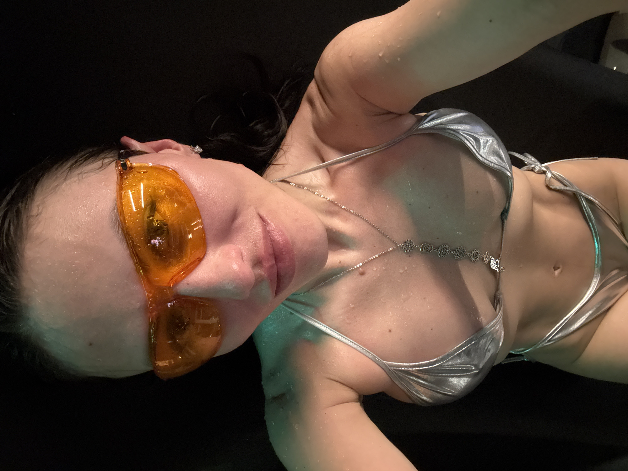 A woman is depicted floating or submerged in a body of dark water, possibly a float tank or pool, in Saint Petersburg, Russia. She is viewed from an elevated perspective, looking towards the camera. She has dark, wet hair and her skin is covered in water droplets, indicating she is wet. She wears a shiny, metallic silver two-piece bikini and large, bright orange, futuristic-style sunglasses. A delicate silver necklace with interconnected circular, floral-like charms hangs around her neck and across her chest. A ring is visible on a finger of her left hand. Subtle green light reflections are noticeable on her skin, particularly on her arm and torso, suggesting ambient or underwater lighting. Her expression is calm and relaxed. No text is visible in the image.