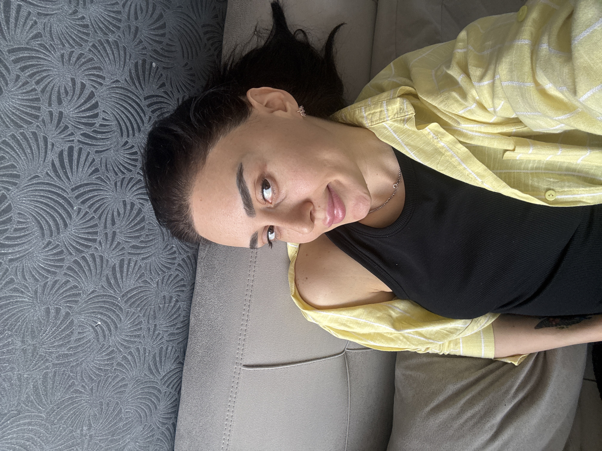 The media file displays a young woman lying on a light gray sofa indoors, photographed from a slightly elevated perspective. She looks upwards towards the camera with a gentle, relaxed smile. Her dark hair is casually styled, appearing somewhat messy around her head. She wears a black ribbed tank top, layered with an unbuttoned, loose-fitting yellow shirt featuring thin white stripes and light yellow buttons. A small, shiny earring is visible in her right ear, and a delicate silver chain necklace adorns her neck. A dark, possibly abstract or floral tattoo is partially visible on her left forearm. The sofa has a subtle texture and stitched detailing. In the background, a dark gray wall is covered with embossed wallpaper showcasing intricate, fan-like or swirling patterns. The lighting suggests natural daylight. There is no visible text in the image. This scene is located in Tbilisi, Georgia.