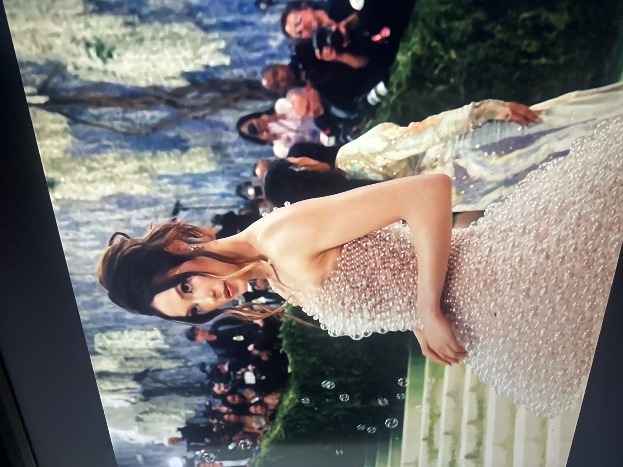 A woman in a white pearl-covered dress stands on stairs. Behind her is a crowd of people and photographers. The background includes what appears to be foliage and an abstract art piece. The setting is not identifiable as Puebla, Mexico from the visual information provided.