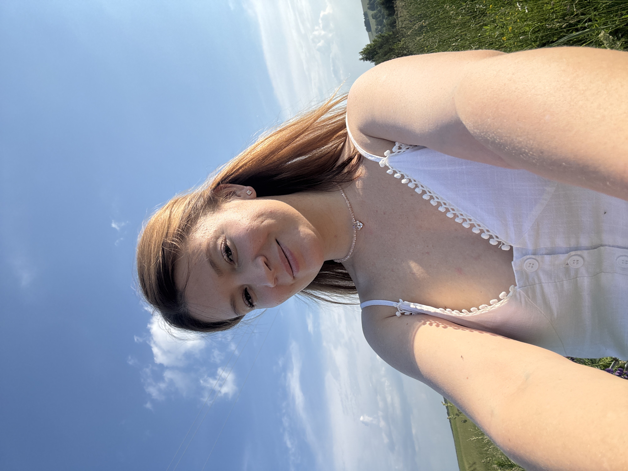 A medium shot portrait shows a young woman with auburn hair taking a selfie outdoors. She is wearing a white sundress with decorative trim and buttons. A delicate necklace with a heart pendant is visible around her neck. Her gaze is directed towards the camera, and she has a slight smile. The background consists of a bright blue sky with scattered white clouds, and a lush green field with tall grass and distant trees. The setting appears to be a rural landscape, possibly in Van'ki, Russia, given the context. The lighting suggests it is daytime, likely late afternoon, due to the warm, golden hue.