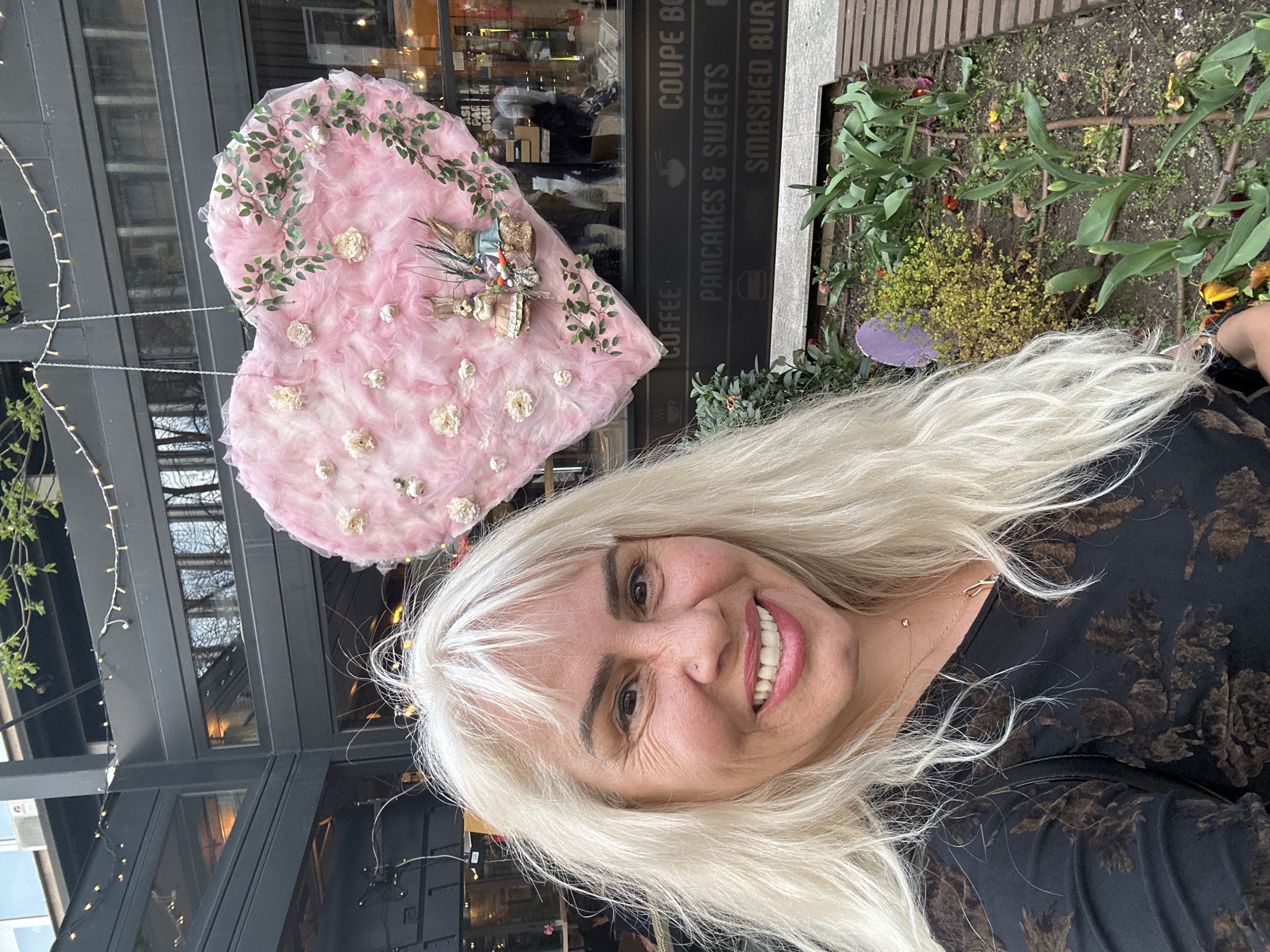 A woman with long white hair is smiling at the camera. Behind her is a large, pink heart-shaped decoration adorned with white flowers and greenery, attached to a pole with string lights. Further in the background, there is a building with large windows and a sign indicating "COFFEE PANCAKES & SWEETS SMASHED BURGER." Next to the building, there is a small garden bed with various green plants and flowers. The photograph appears to be taken outdoors during the daytime. Given the sign's reference to food and the architectural style of the building, the location may be near a restaurant or cafe. Without additional clues, the specific location in Sofia, Bulgaria cannot be definitively determined.
