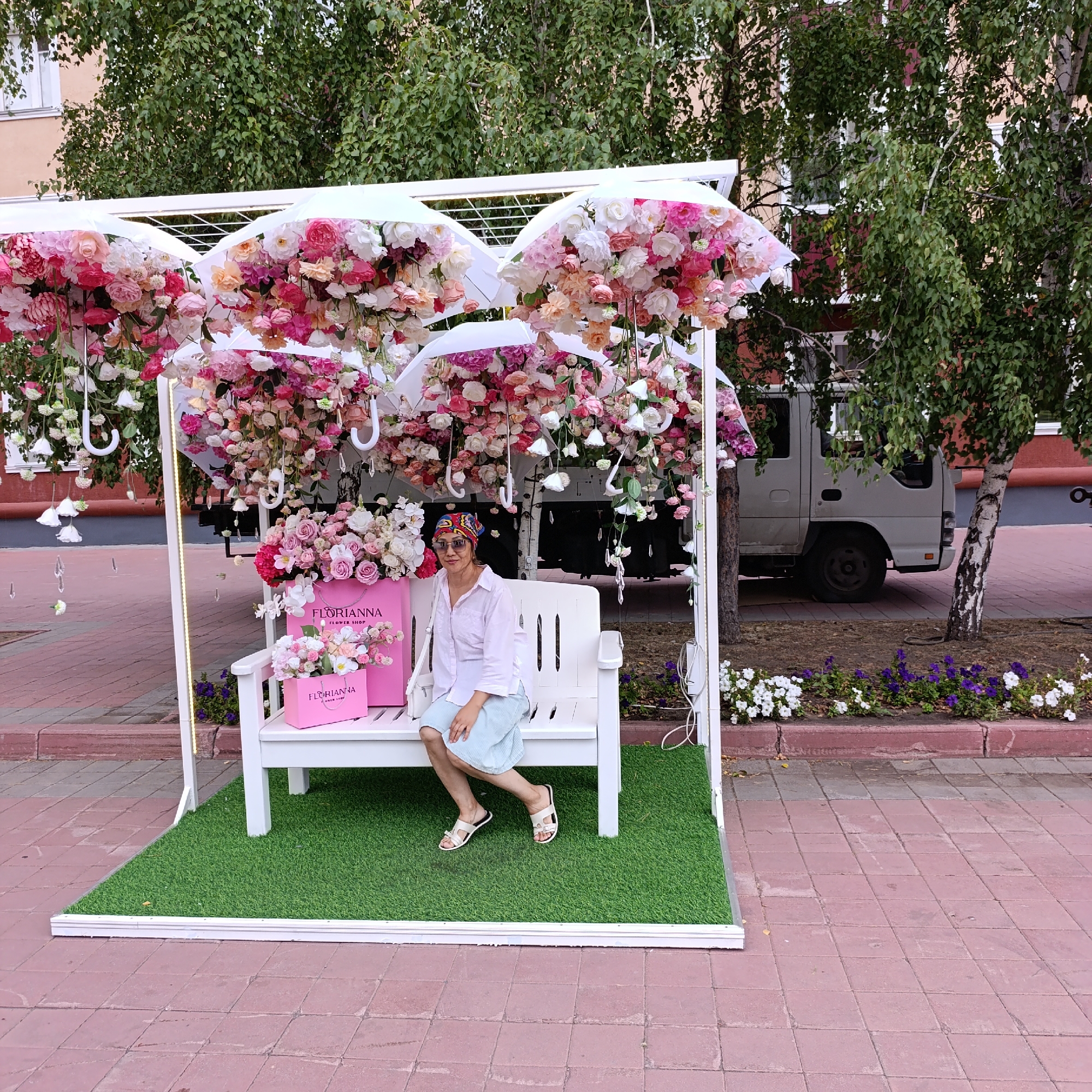 In the city of Kostanay, Kazakhstan, on a paved walkway lined with flowering plants, a woman sits on a white bench. The bench is situated on a patch of artificial green grass, in front of an artistic display. This display features a white frame from which several white umbrellas are suspended, adorned with abundant clusters of pink, white, and peach-colored artificial flowers, creating a cascading floral effect. Below the umbrellas, also on the bench, are two pink paper shopping bags with "FLORIANNA FLOWER SHOP" printed on them, and a smaller bouquet of flowers. The woman, wearing sunglasses, a patterned headscarf, a light pink collared shirt, and a pale blue skirt, is casually posed with her legs crossed. Behind the display, a light-colored truck is partially visible, parked near trees with green foliage. The overall scene suggests an outdoor decorative setup, possibly for an event or a commercial establishment.