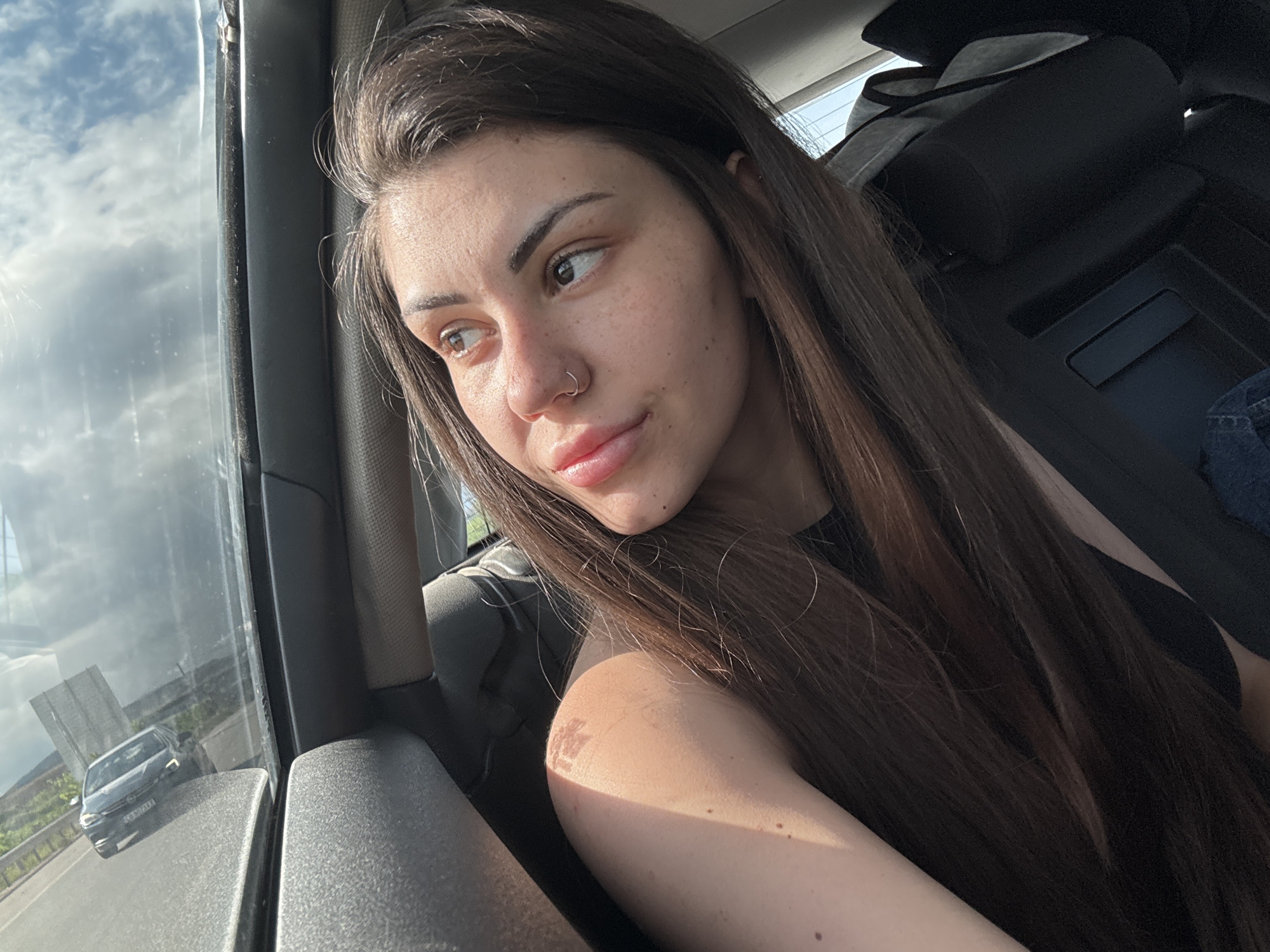 A young woman with long, dark brown hair and a nose piercing is seen from the chest up, seated inside a car and looking out of the passenger side window. Her expression is calm and observant, and bright sunlight illuminates the right side of her face and arm, highlighting freckles and a possible faint mark on her bicep. She wears a dark top.
Through the car window, a bright daytime sky with scattered clouds is visible. The reflection on the window prominently features a dark-colored car, possibly a Volkswagen Golf, driving on a road or highway alongside a guardrail. The reflected car's headlights are on, and a partial license plate, "M117AA" with "CJ" prefix, is discernible, indicating a Romanian vehicle. Distant hills or elevated terrain are faintly visible beyond the road. The setting suggests the woman is a passenger during a car journey in or around Hrabarsko, Bulgaria.