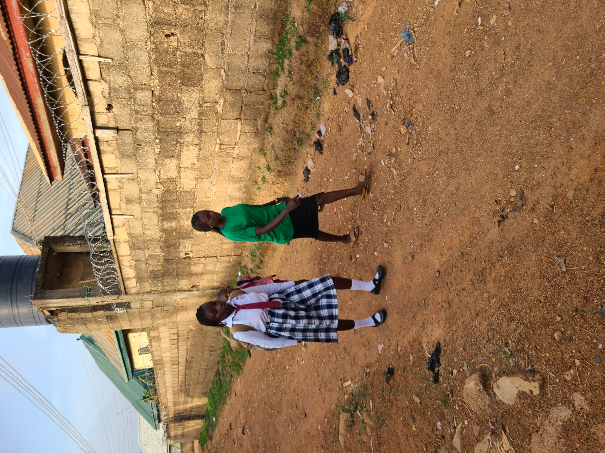 Two young African females are on a dirt ground in Jos, Nigeria. One female wears a green top with a black skirt and the other wears a white shirt with a plaid skirt, a backpack and a red tie, presumably a school uniform. The ground is scattered with trash. A cinder block wall is to the left with barbed wire on top. A building with a green roof is visible.