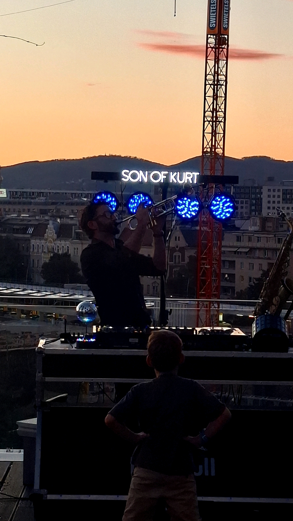 In Vienna, Austria, at dusk, a man plays a trumpet on a rooftop stage, illuminated by blue stage lights. Behind him, a neon sign reads "SON OF KURT". A child stands with hands on hips, facing away from the camera, towards the stage. A construction crane with "SWIETEL" written on it is visible in the background, along with city buildings and a hazy orange sky. A saxophone rests on a stand to the right of the stage.